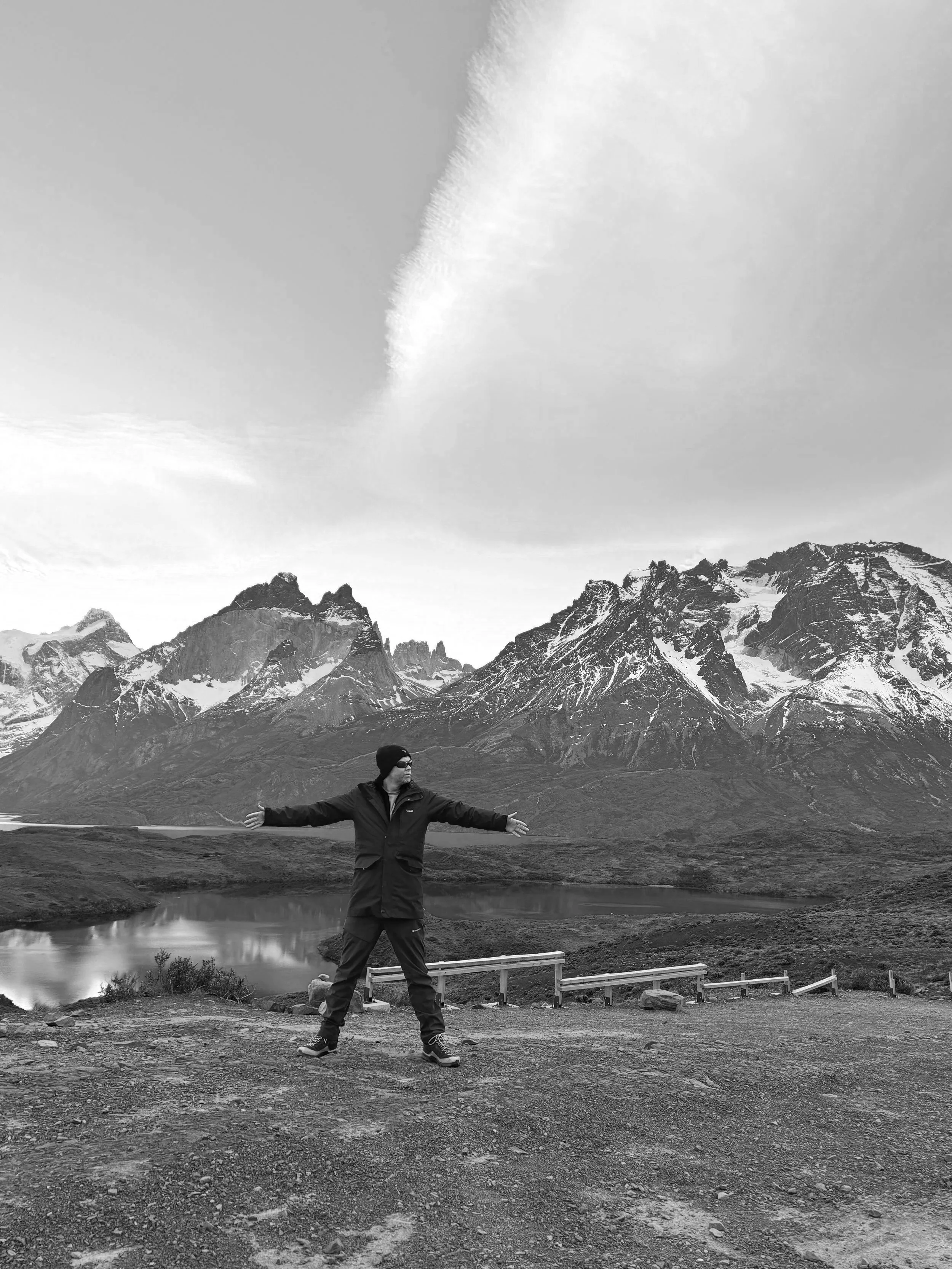 A person standing with arms outstretched in front of a lake and snow-capped mountains under a blue sky with large white clouds.