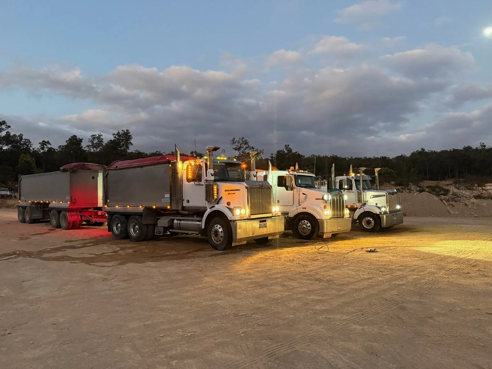 Three large semi-trucks parked on a dirt area during dusk with a partly cloudy sky in the background.
