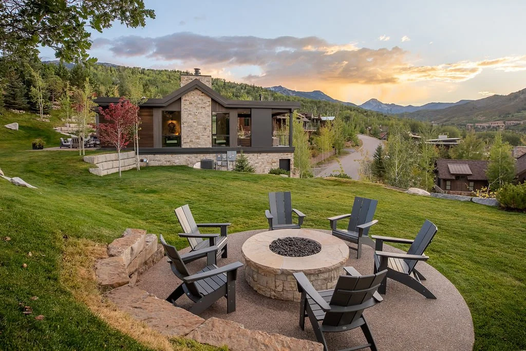 A backyard with a fire pit surrounded by six chairs, green lawn, trees, and a house in the background, with mountains in the distance at sunset.