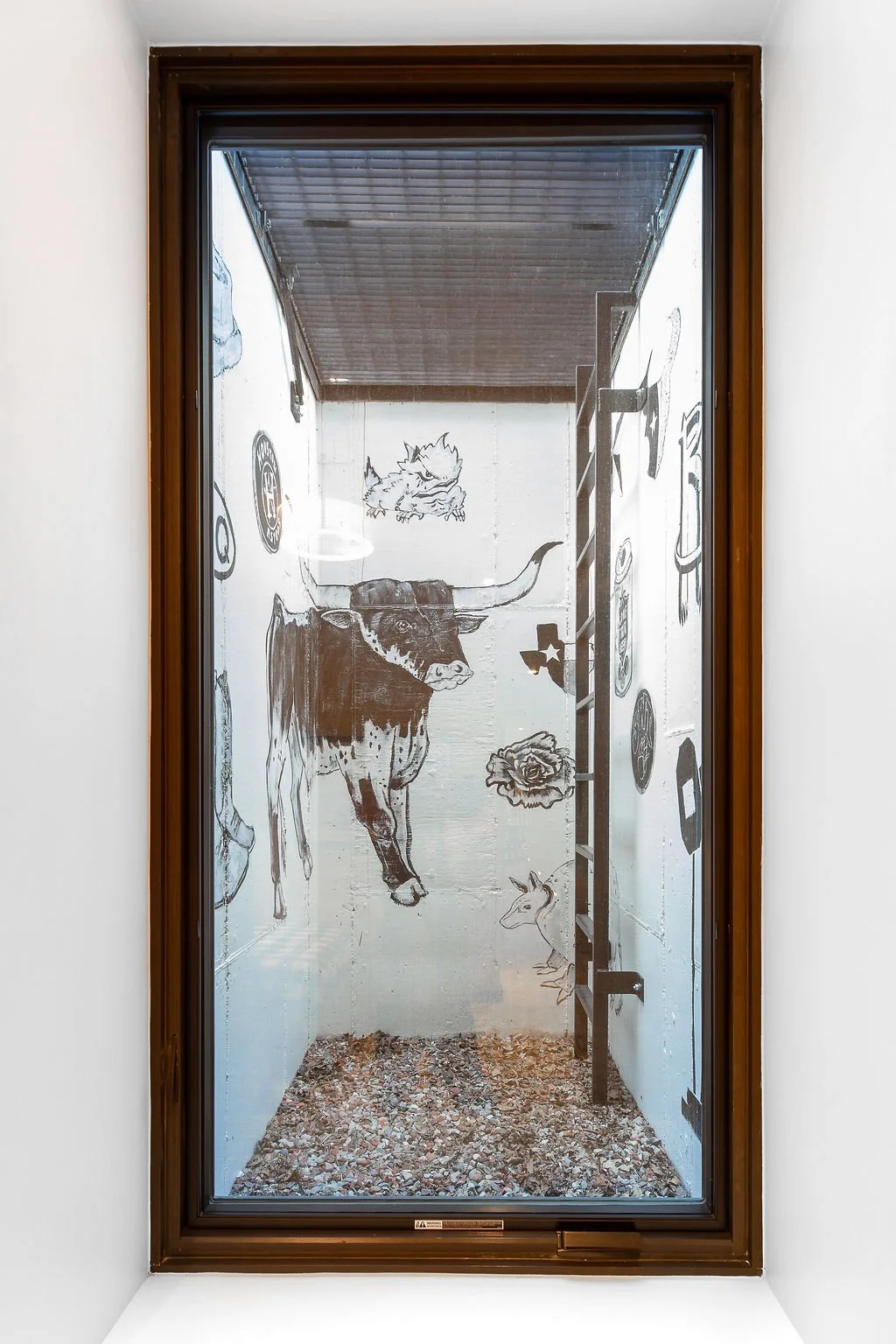 Small enclosed space with white walls decorated with black line drawings of animals and objects, including a longhorn steer, a horse, a cat, a dog, a rose, a star, and various symbols, viewed through a window.