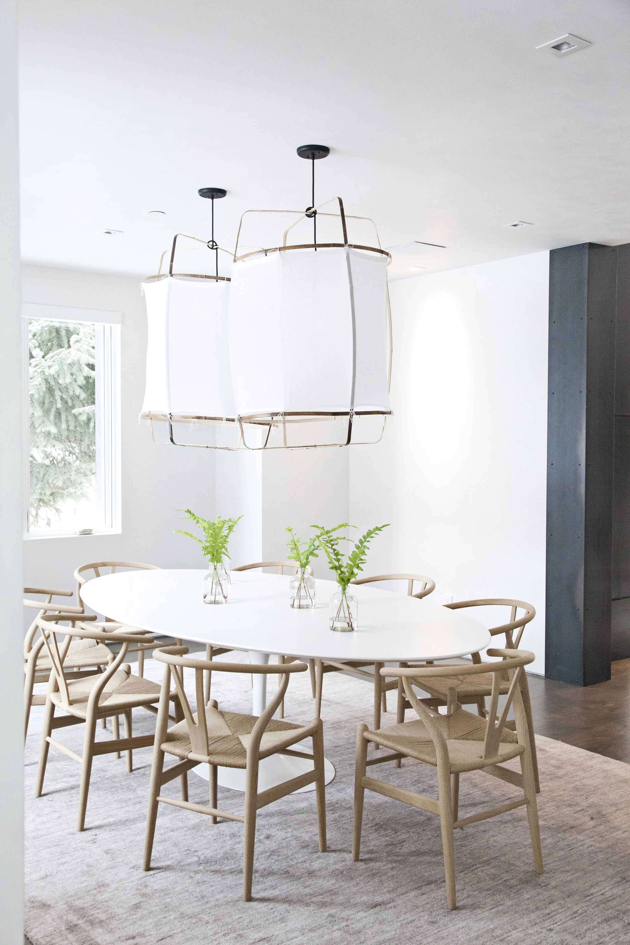 Modern dining room with an oval white table, six wooden chairs with woven seats, three glass vases with green fern plants on the table, a large white fabric and wood chandelier overhead, a window showing outdoor greenery, and a wooden floor with a li
