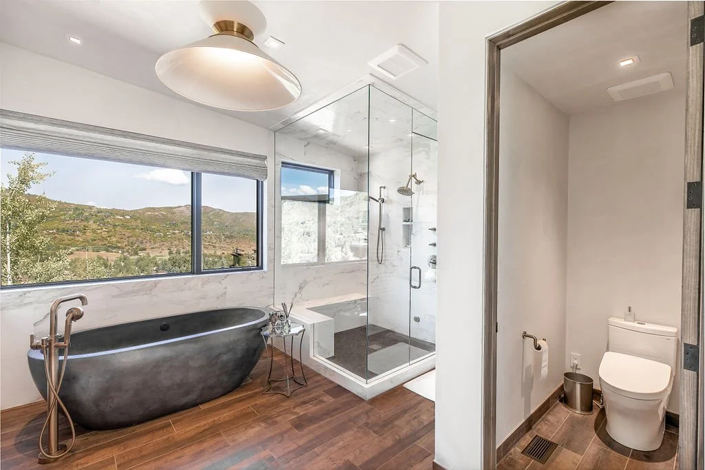 Modern bathroom with a black freestanding bathtub, large window with mountain view, glass shower enclosure, and separate toilet area.