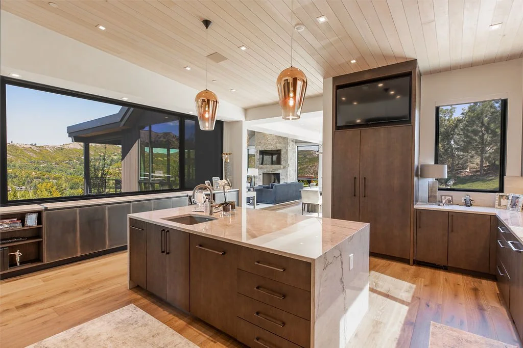 Modern kitchen with wooden cabinets, a marble island, large windows showing a scenic outdoor landscape, and a view of the living room with a fireplace.