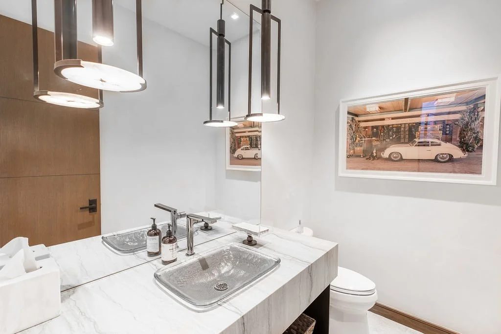 Bathroom with dual sinks, modern light fixtures, white walls, and a framed photo of a vintage white car on the wall.