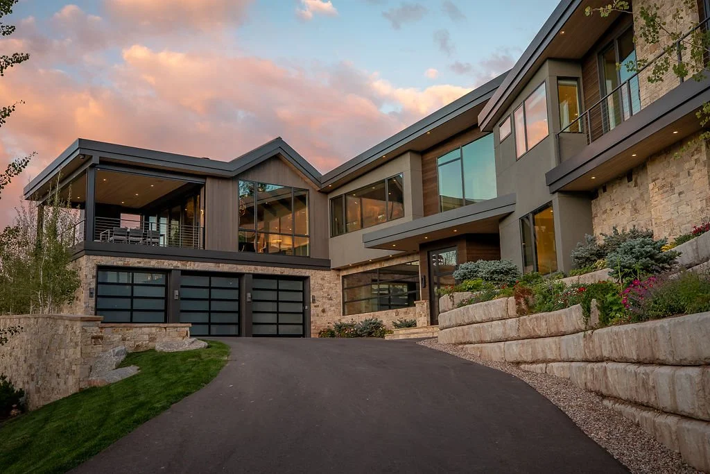 A modern multi-story house with large glass windows, stone and wood exterior, and a landscaped sloped driveway under a sunset sky.