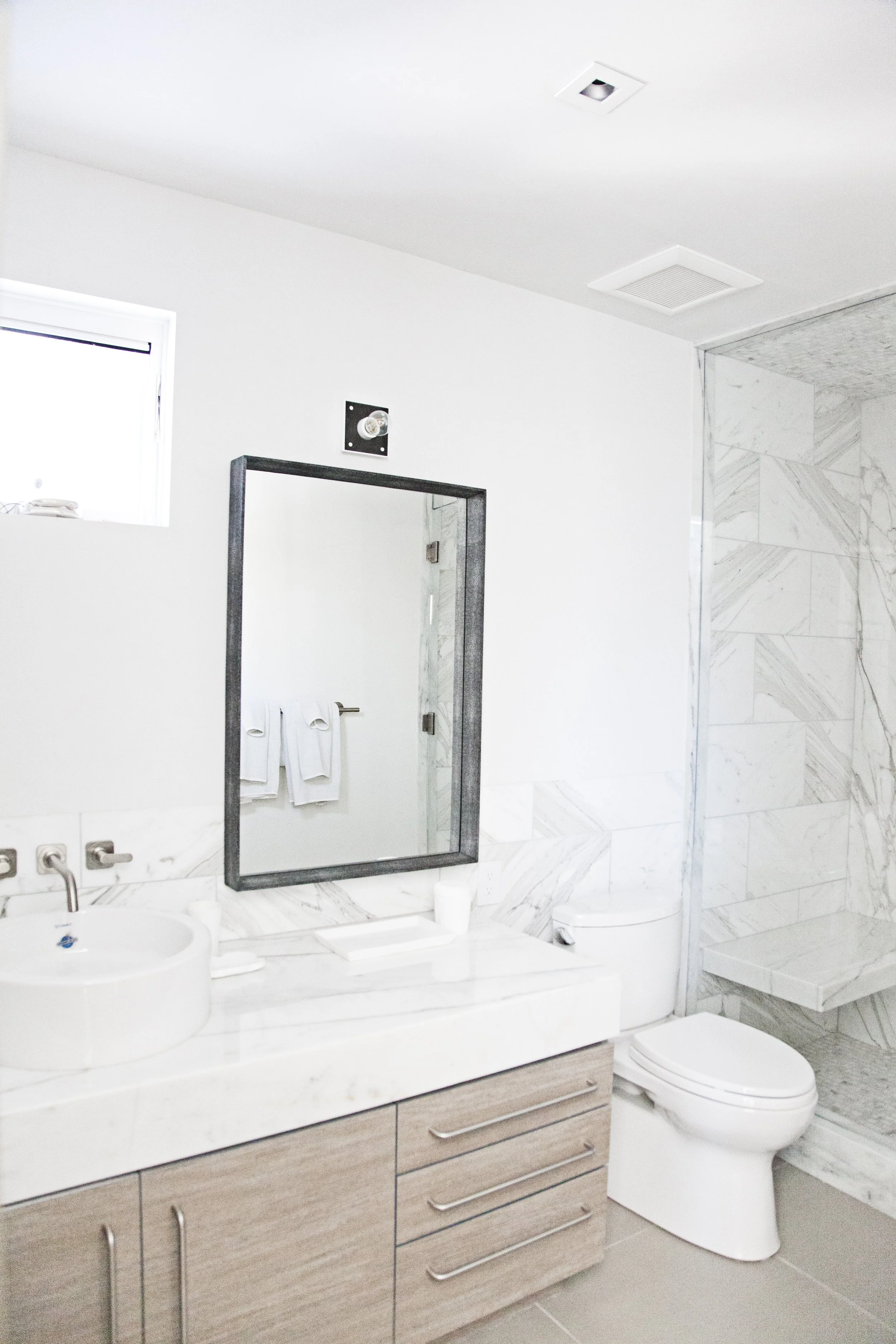 Modern bathroom with white walls, a window, a rectangular mirror with a black frame, two towels hanging, a white countertop with a small sink, a toothbrush holder, a toilet, and a walk-in shower with marble tiles.
