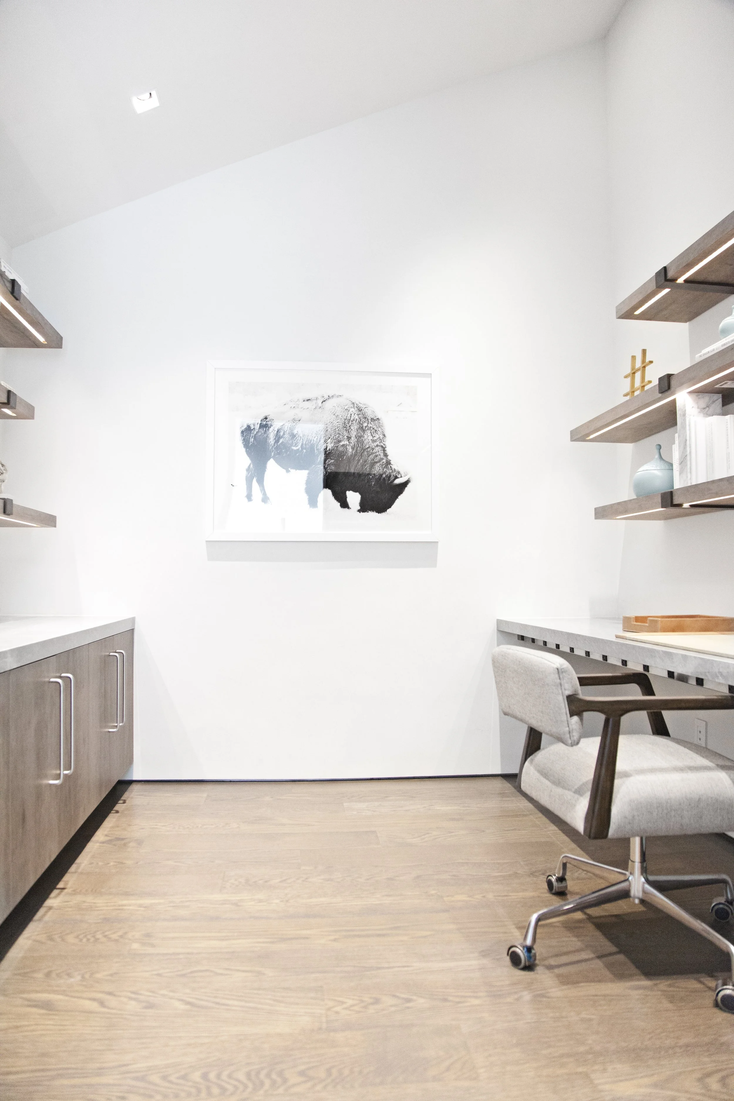 Modern home office with wooden floor, white walls, framed bison artwork, floating shelves with vases and books, desk, and cushioned office chair.