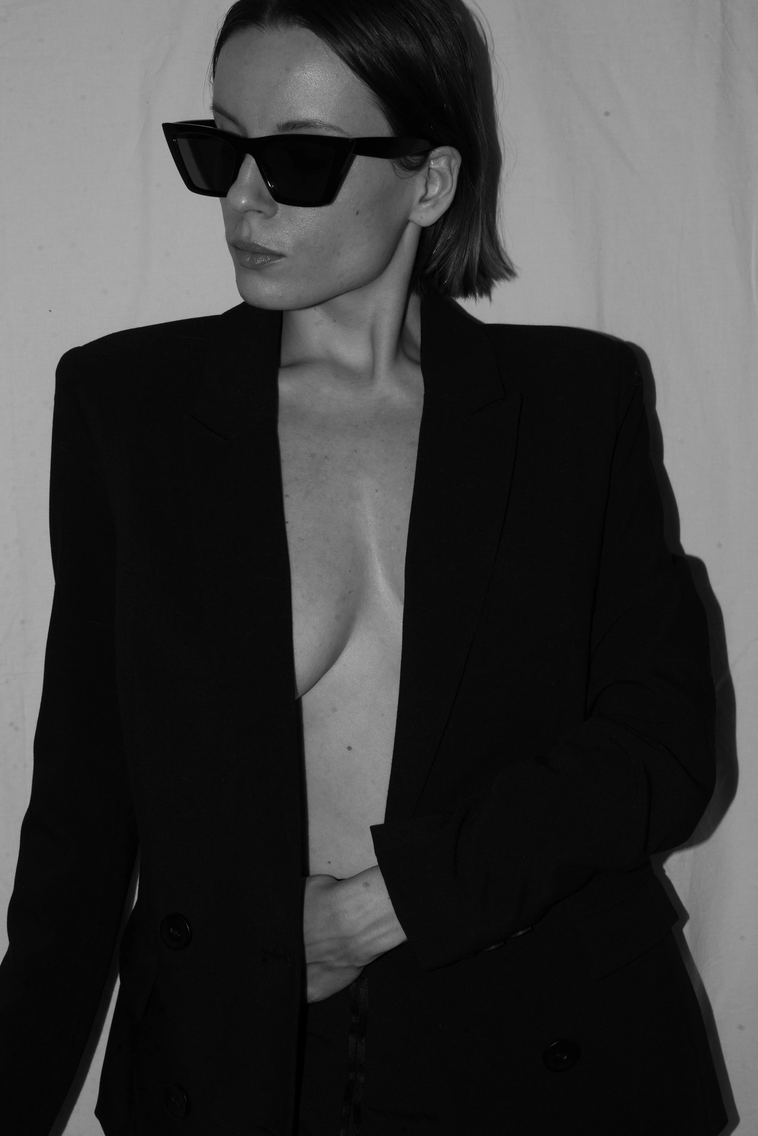 A woman wearing black sunglasses and a black blazer, topless underneath, with her hand on her stomach, standing against a plain background.