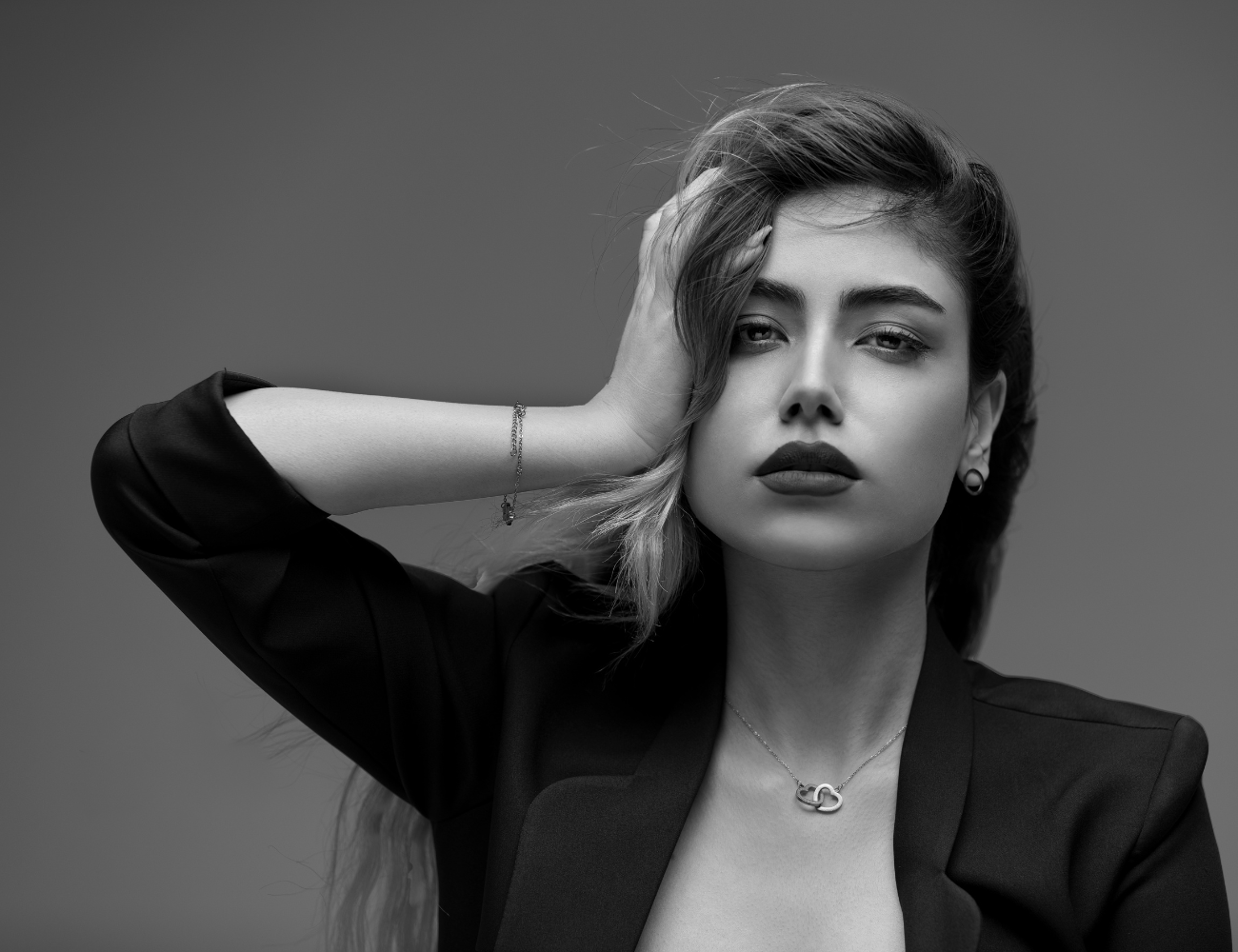 A woman with wavy hair and bold lipstick posing with her left hand on her head, wearing a dark blazer, a necklace, and earrings, in a black and white studio portrait.
