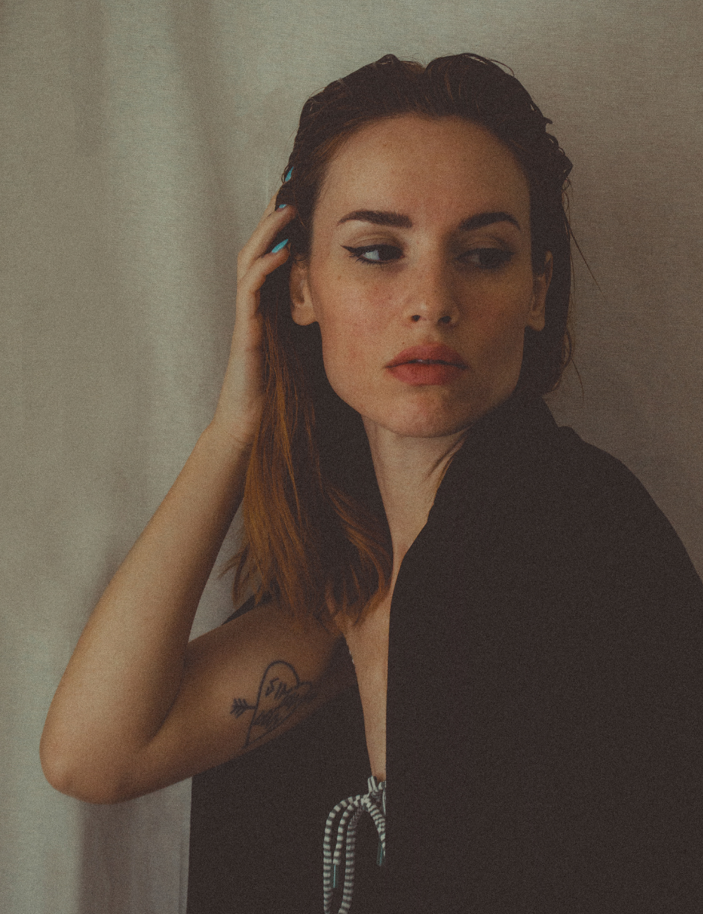 A young woman with reddish-brown hair, wearing black clothing, is touching her hair with one hand and looking slightly to the side against a plain textured background.
