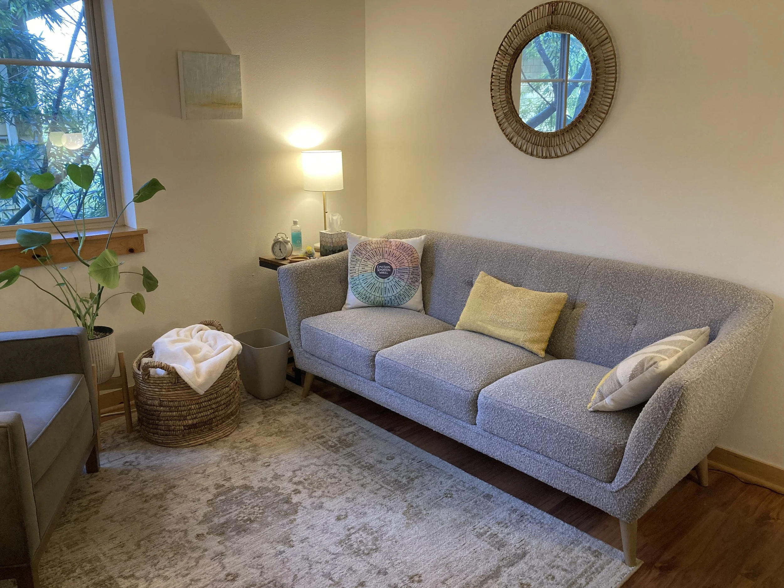 A cozy living room with a gray sofa adorned with colorful pillows, a window with greenery outside, a round wicker mirror on the wall, and a side table with a lamp, clock, and tissues.