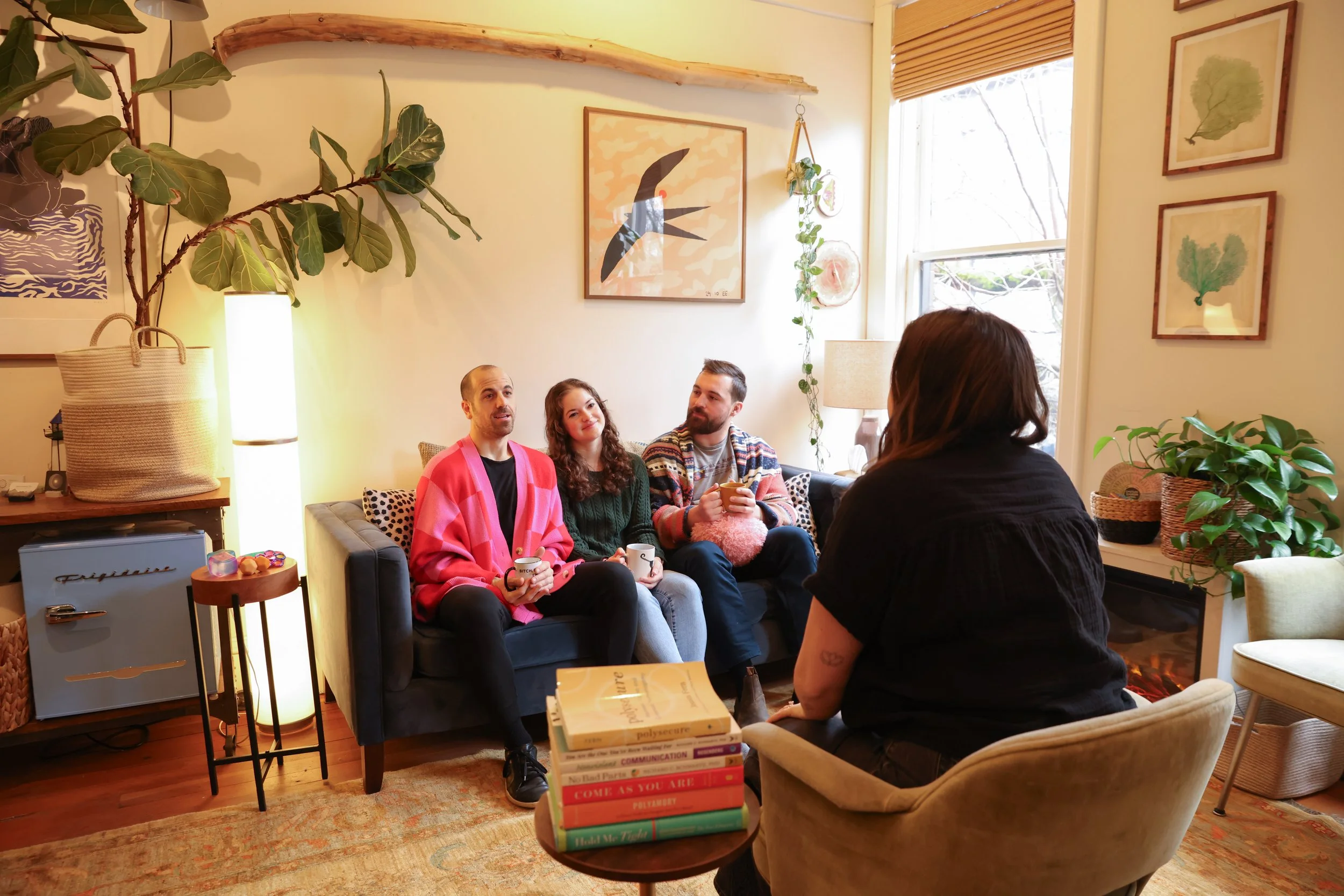 Four people having a conversation in a cozy living room, three sitting on a couch and one on a chair, with plants and artwork on the walls.