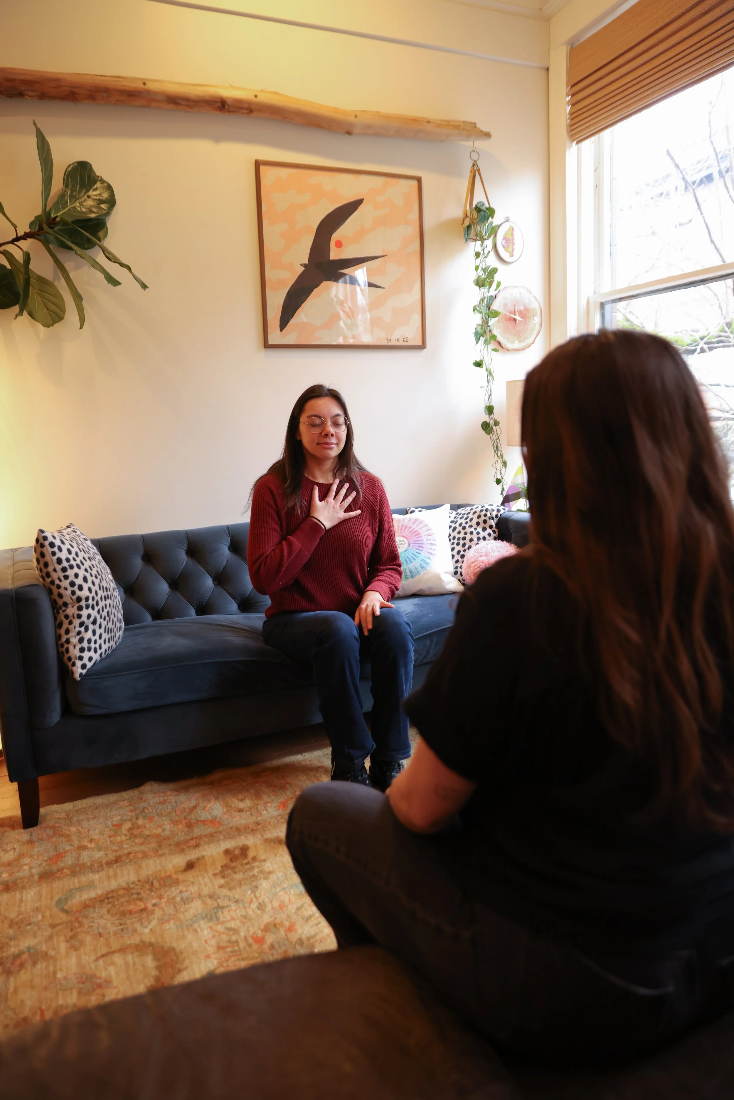Two women sitting in a living room having a conversation, one woman is on a dark couch with a hand on her chest, the other is sitting with her back to the camera. The room is decorated with plants, pillows, and artwork, with a window letting in natural light.