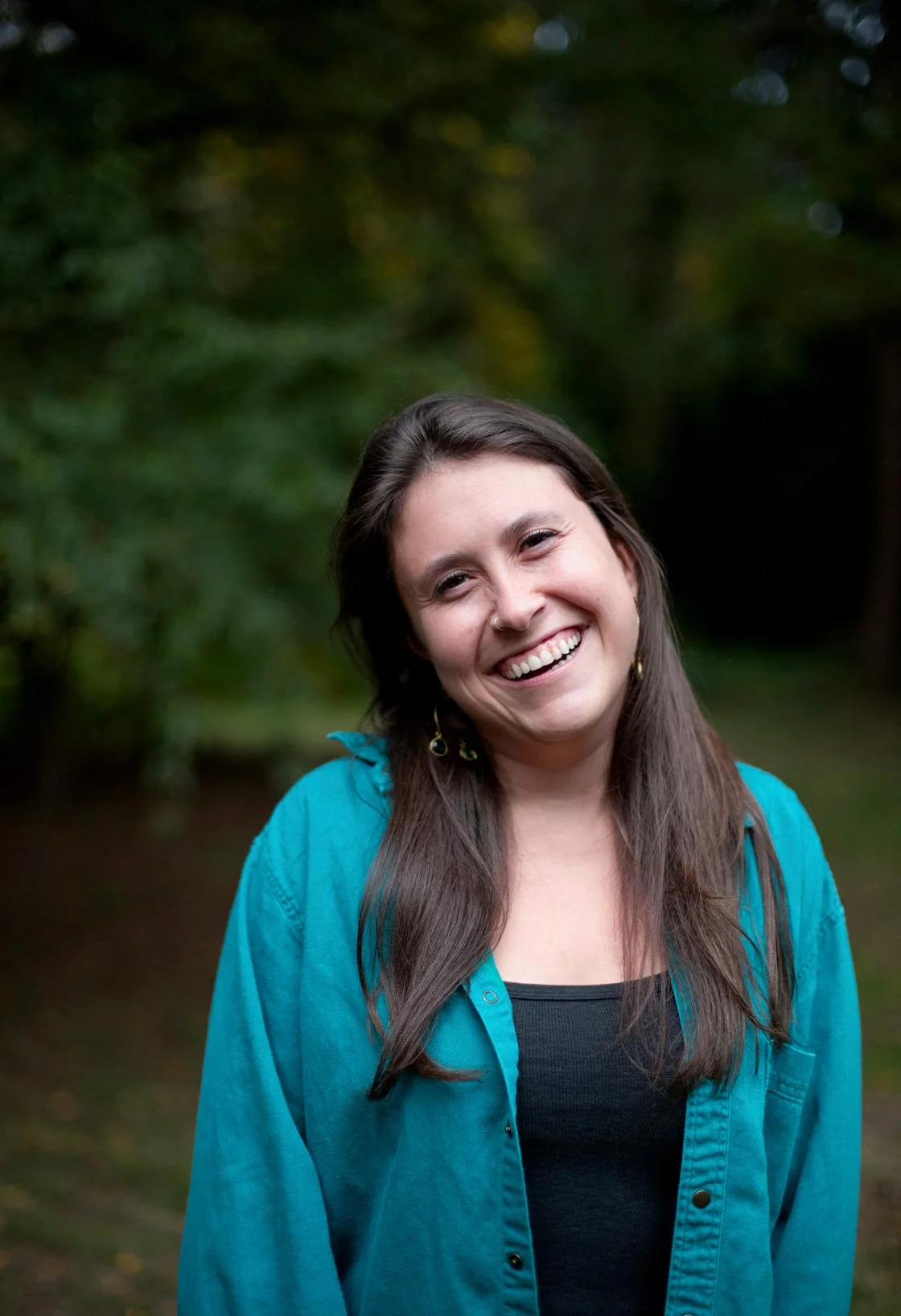 A woman with long brown hair and a nose ring smiling outdoors, wearing a black top and a blue jacket.
