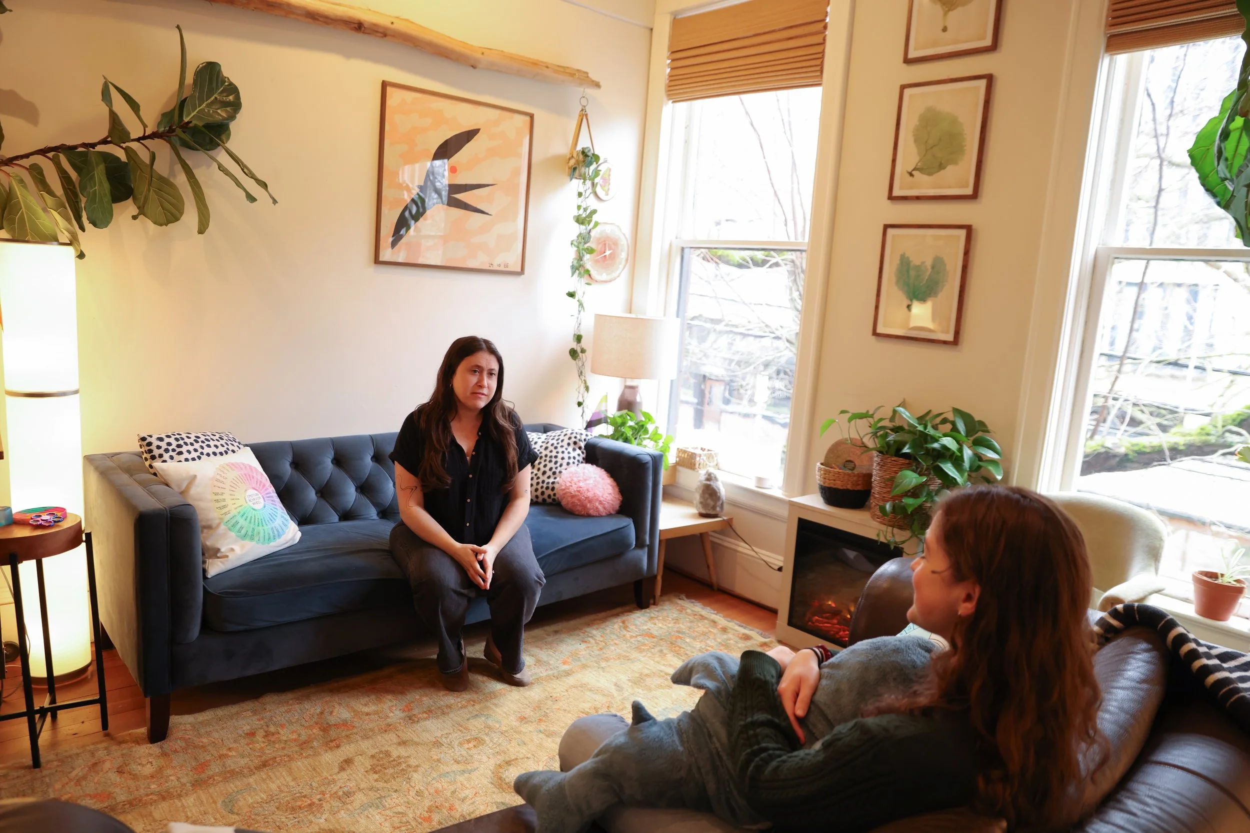Two women sitting and talking in a cozy living room with large windows, artwork, plants, and a fireplace.