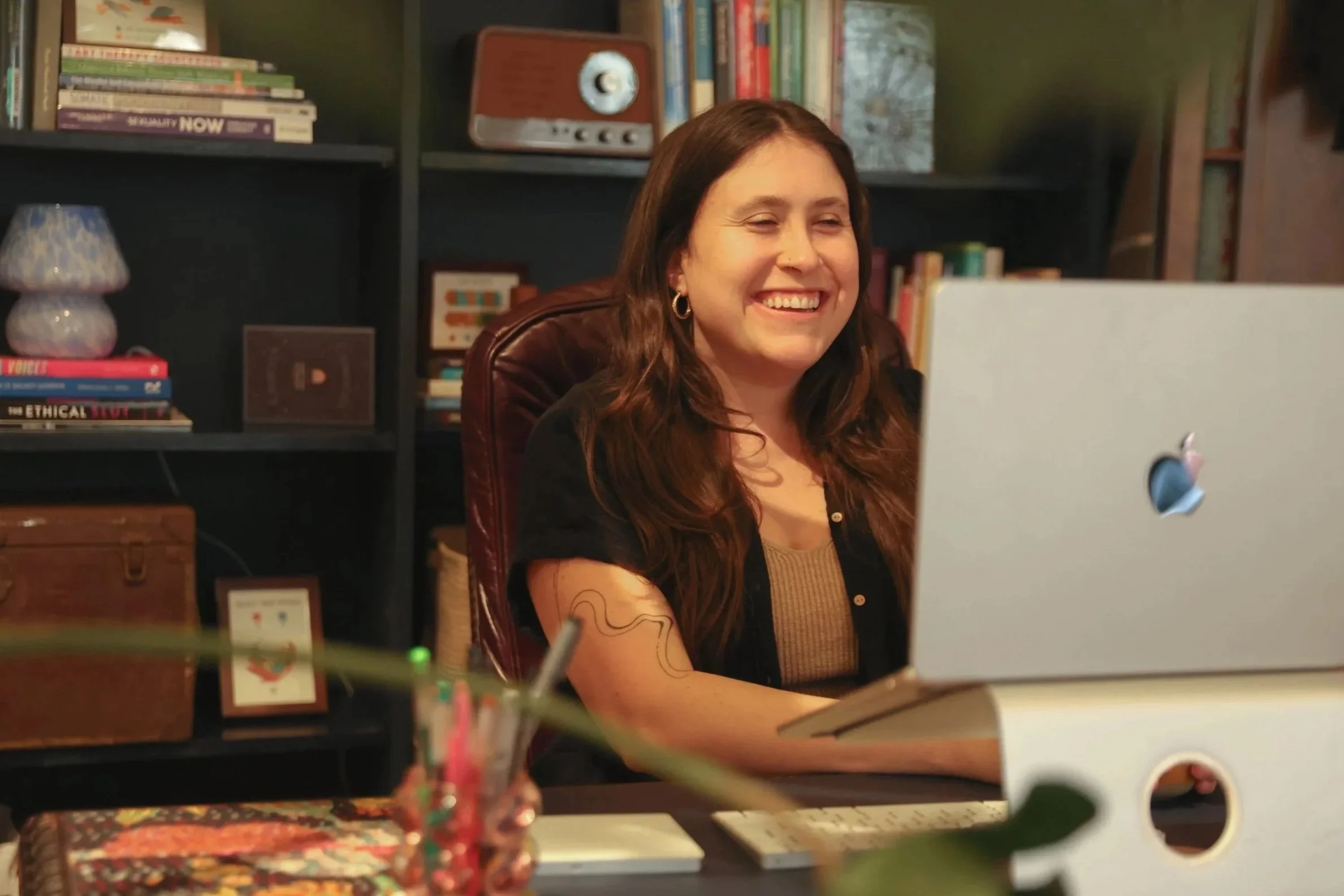 Two women sitting on a gray couch looking at a laptop with a colorful geometric pattern. One woman wearing glasses and a light blue shirt, the other holding a yellow mug and wearing a green sweater. There are decorative pillows behind them and a rainbow art piece on the windowsill.