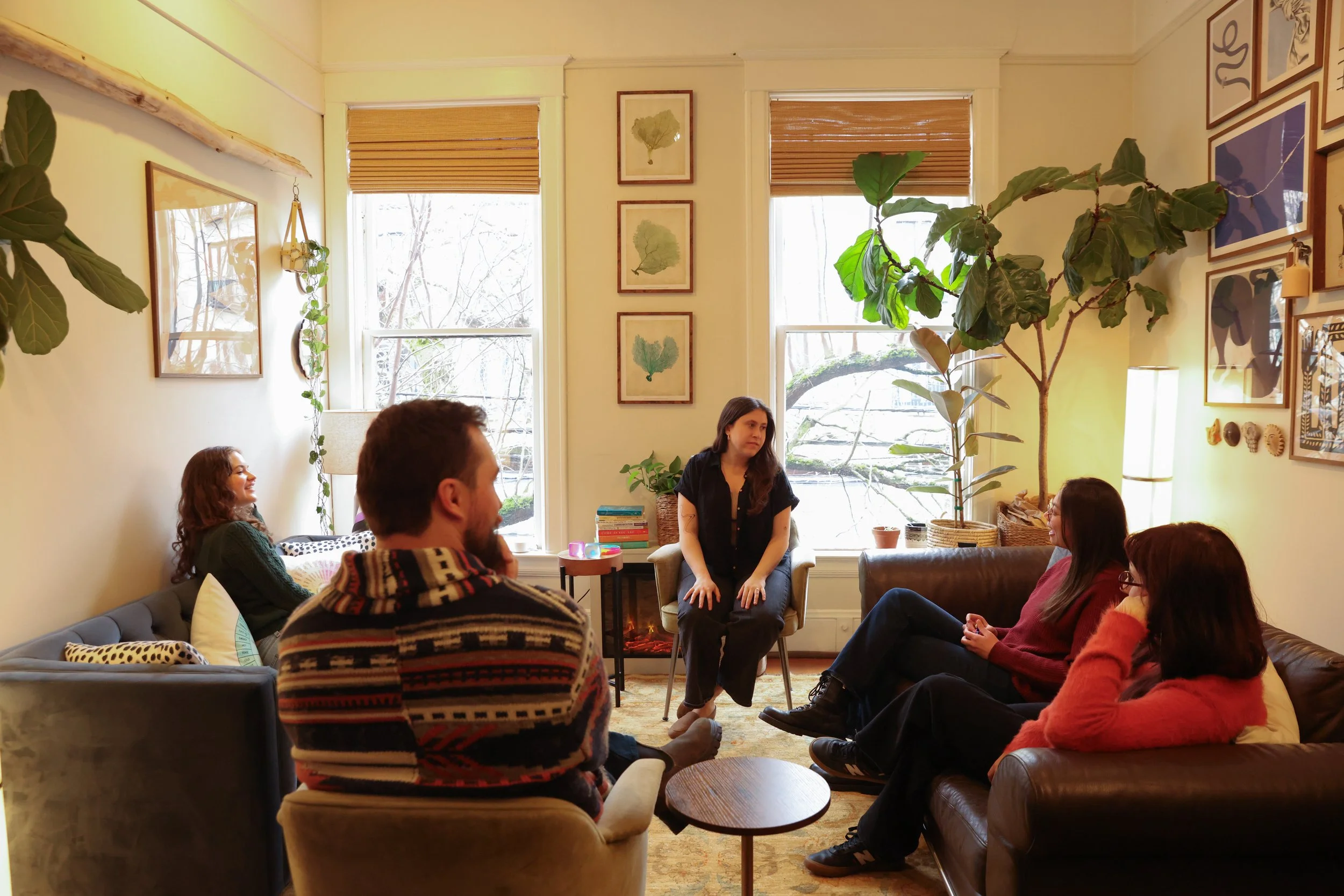 People attending a group therapy session in a cozy living room, seated on sofas and chairs, with a woman leading the session in the center. The room has large windows, plants, framed artwork, and a bookshelf.