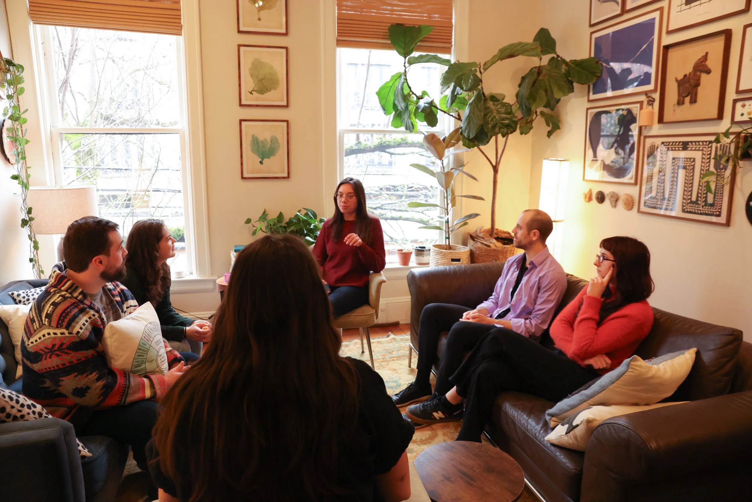 A woman giving a presentation or speech to five seated people in a cozy, art-decorated living room with large windows and indoor plants.