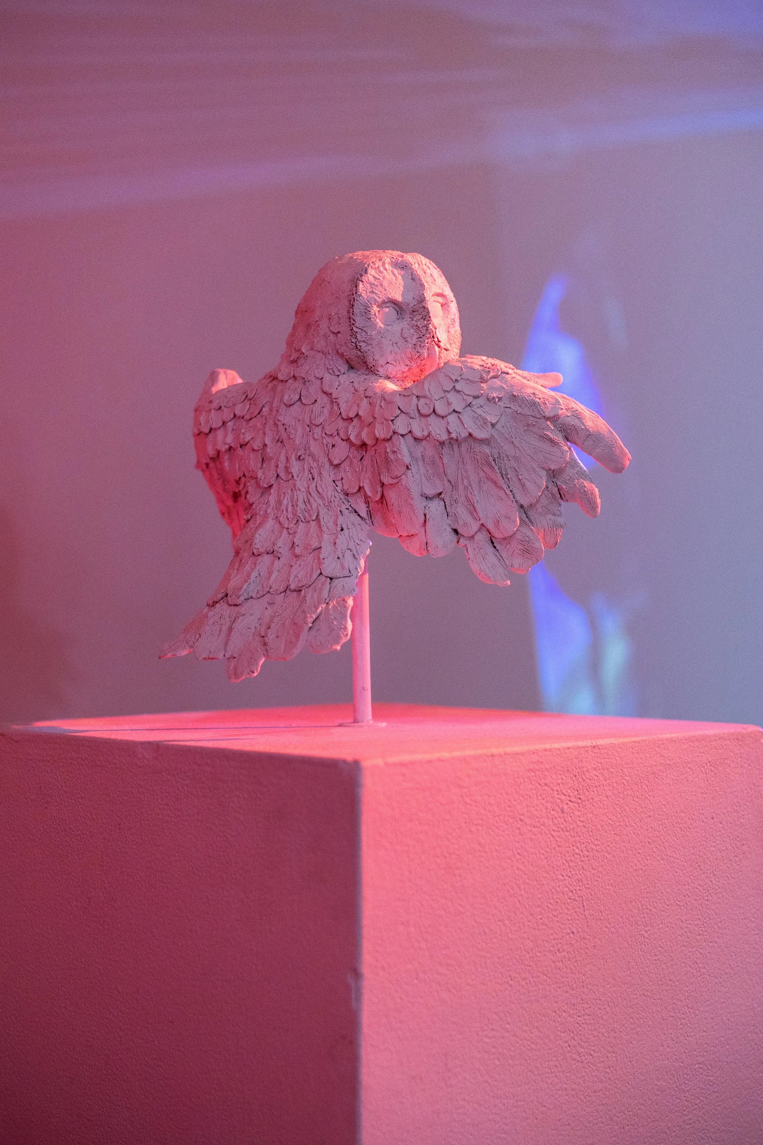 A sculpture of an owl with textured feathers on a pink pedestal, lit with pink and purple lighting, with blurred blue and purple background elements.