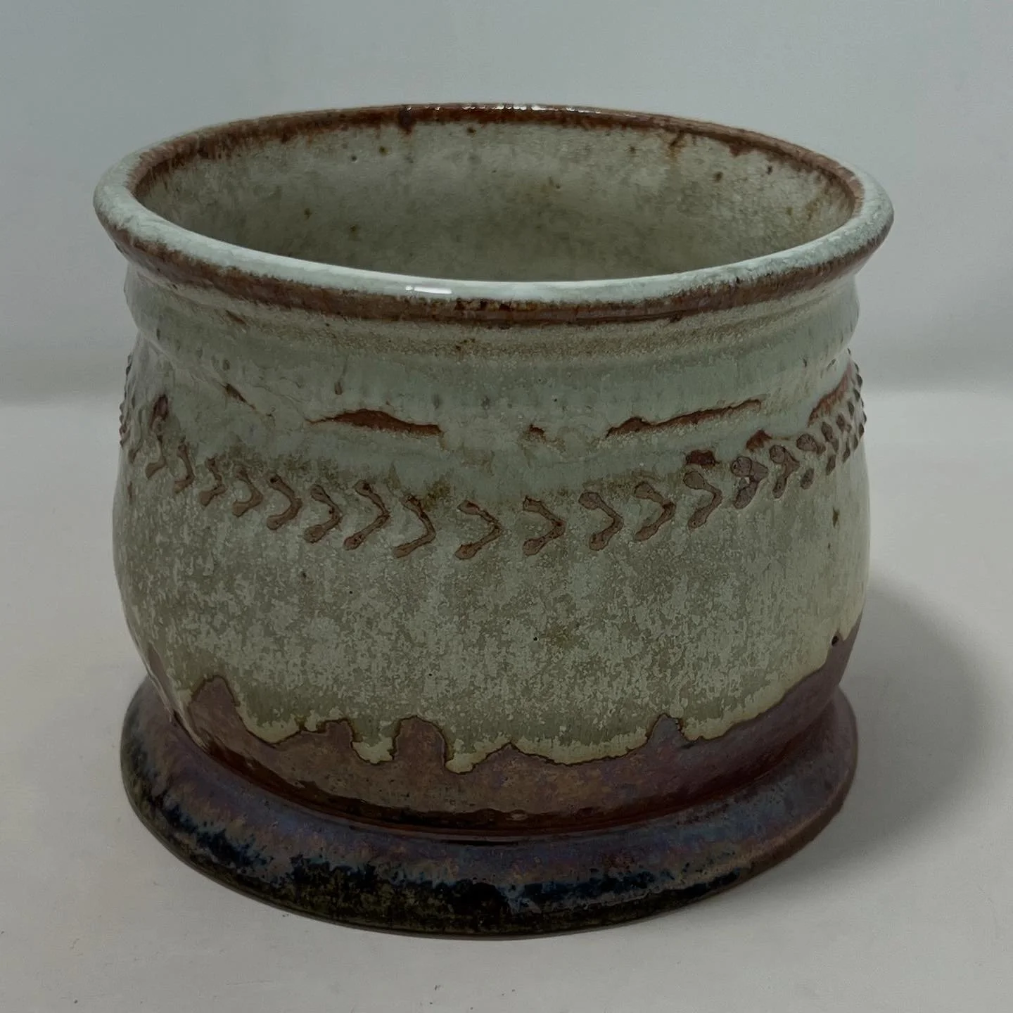 A ceramic pot with a decorative pattern around its middle, glaze with a weathered, rustic appearance, sitting on a white surface.