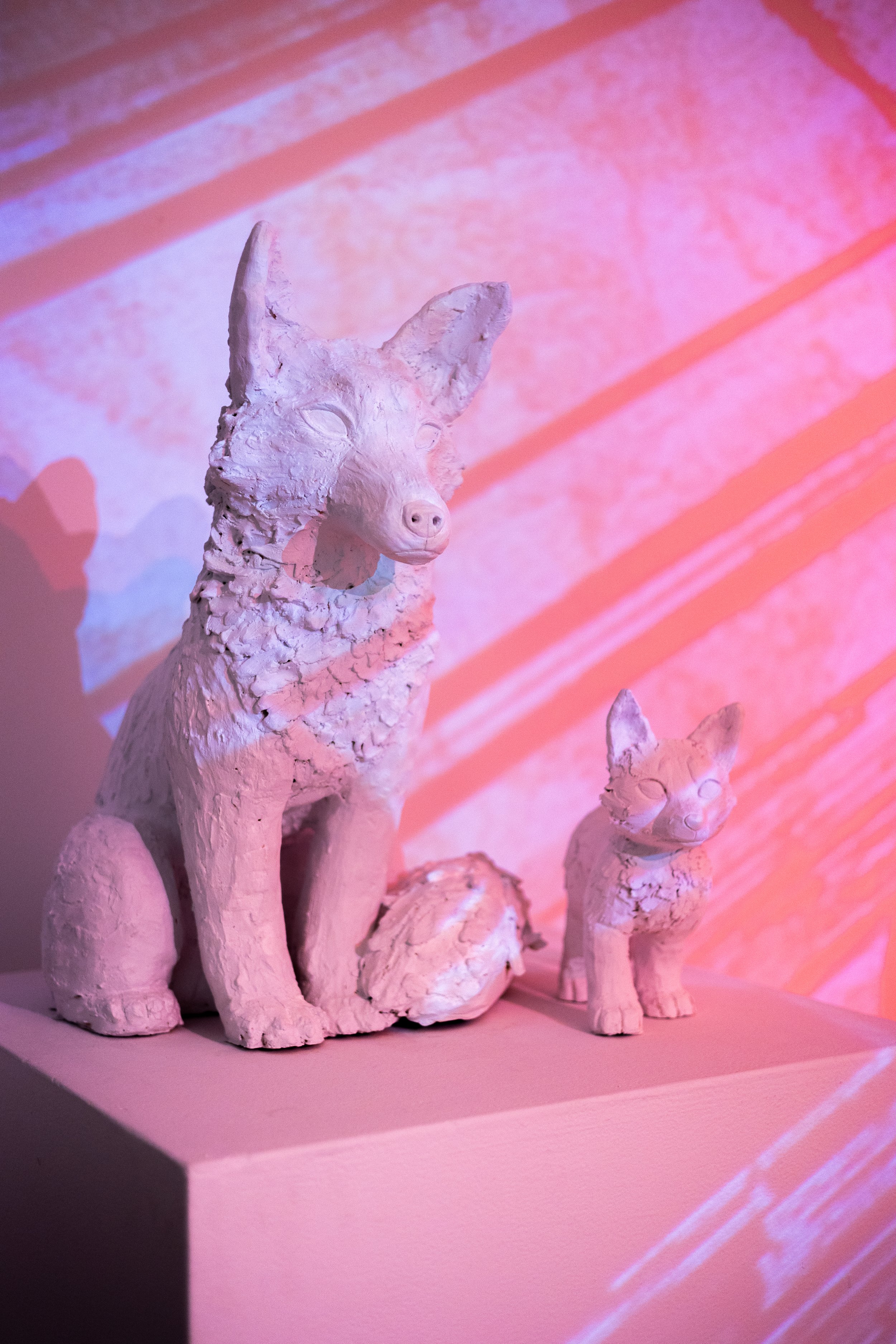 White ceramic sculptures of a dog, a cat, and smaller animals, displayed on a pink platform against a pink and orange abstract background.