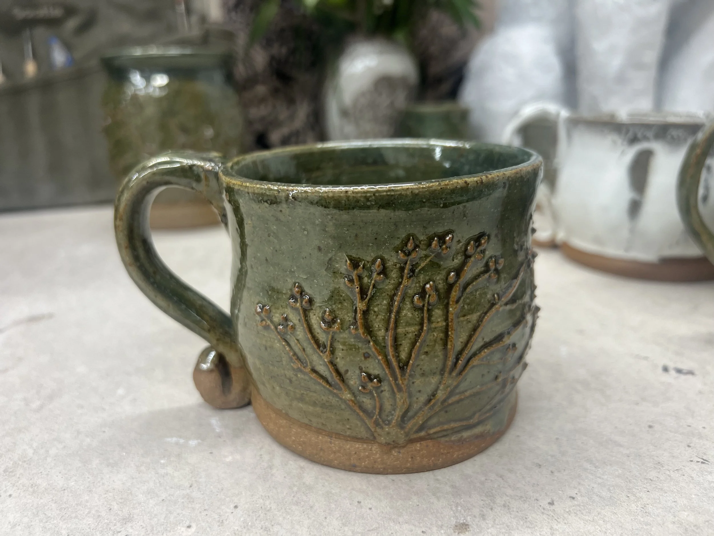 A handmade ceramic mug with a green glaze and a decorative design of raised branches and buds. The mug sits on a light-colored surface with other cups blurred in the background.