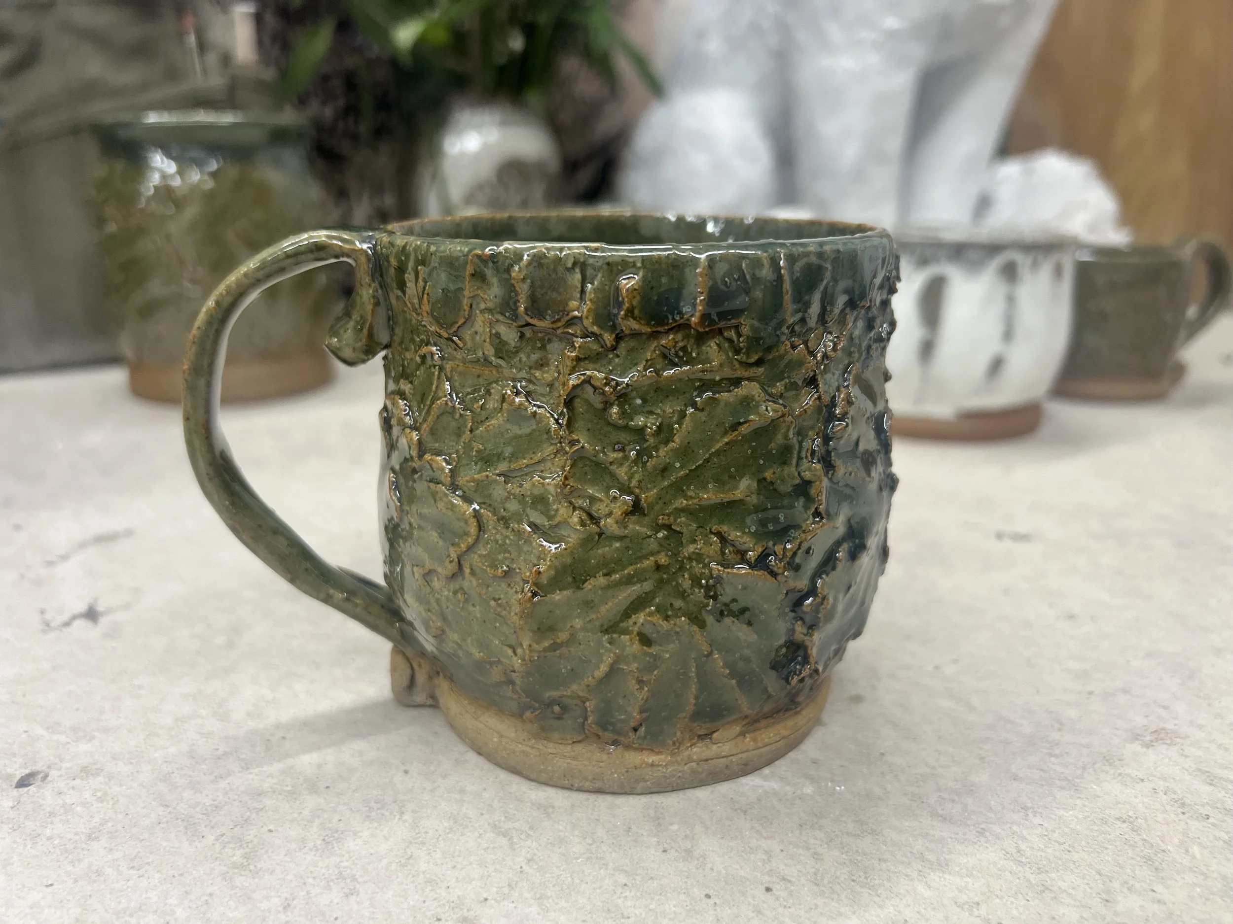 A ceramic cup with a green glaze and a textured, uneven surface, sitting on a white surface with other blurred ceramic items in the background.