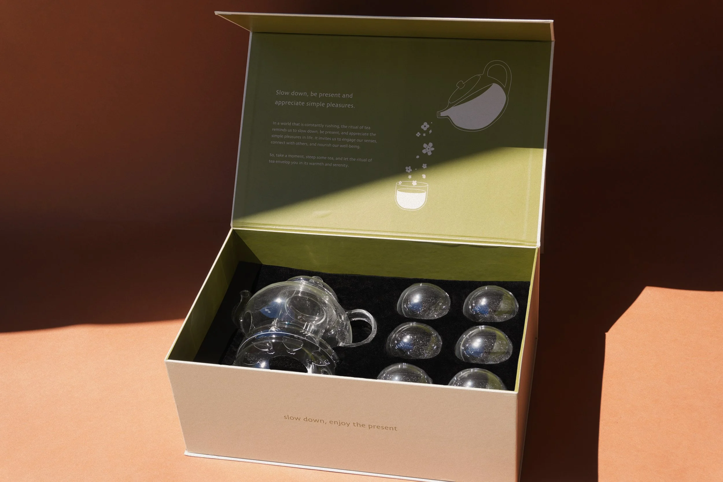 Glass teapot and cups set in a beige box with green interior lid featuring a teapot illustration and text about enjoying simple pleasures.