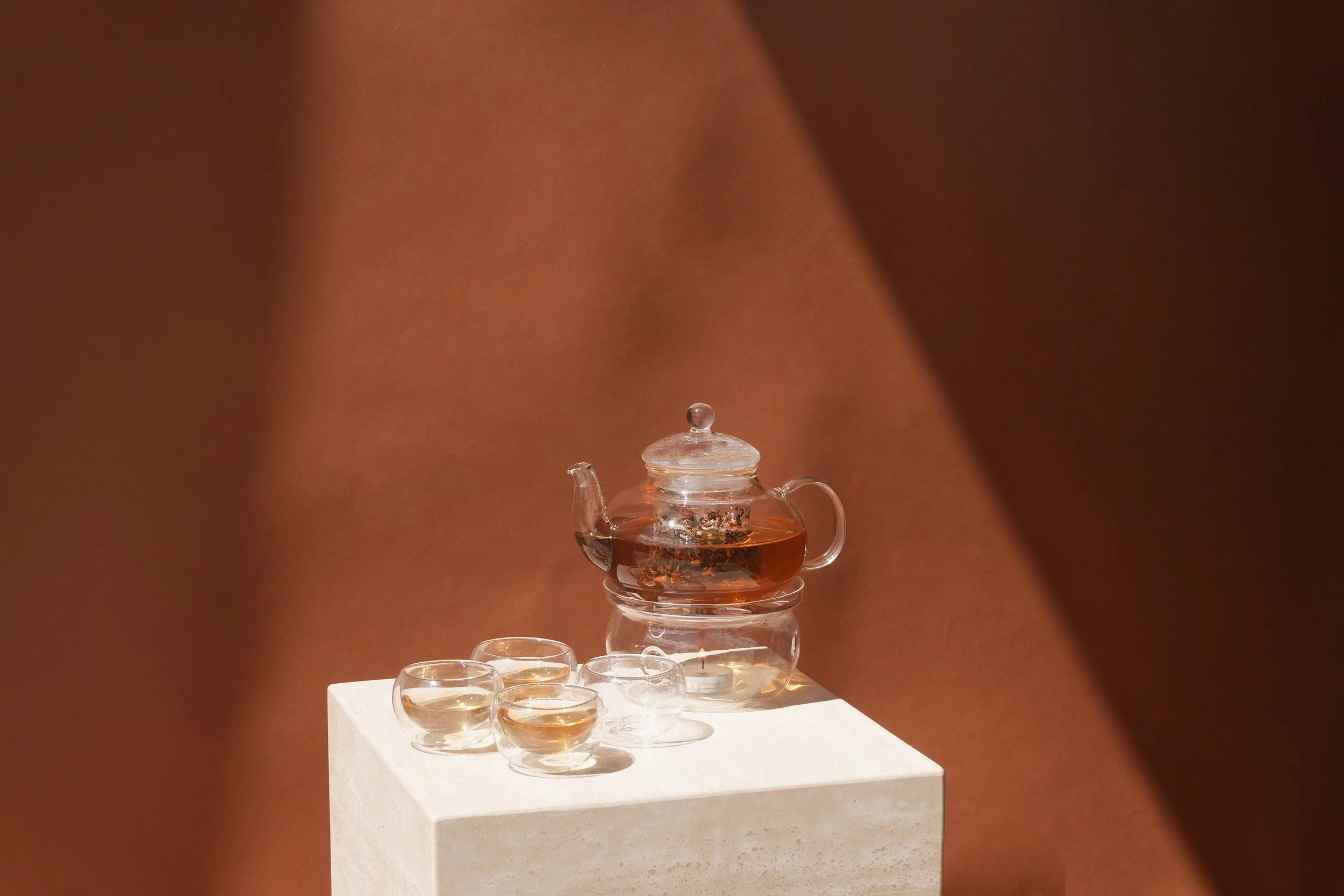 Glass teapot with tea and four glass cups on a white pedestal against a brown background.