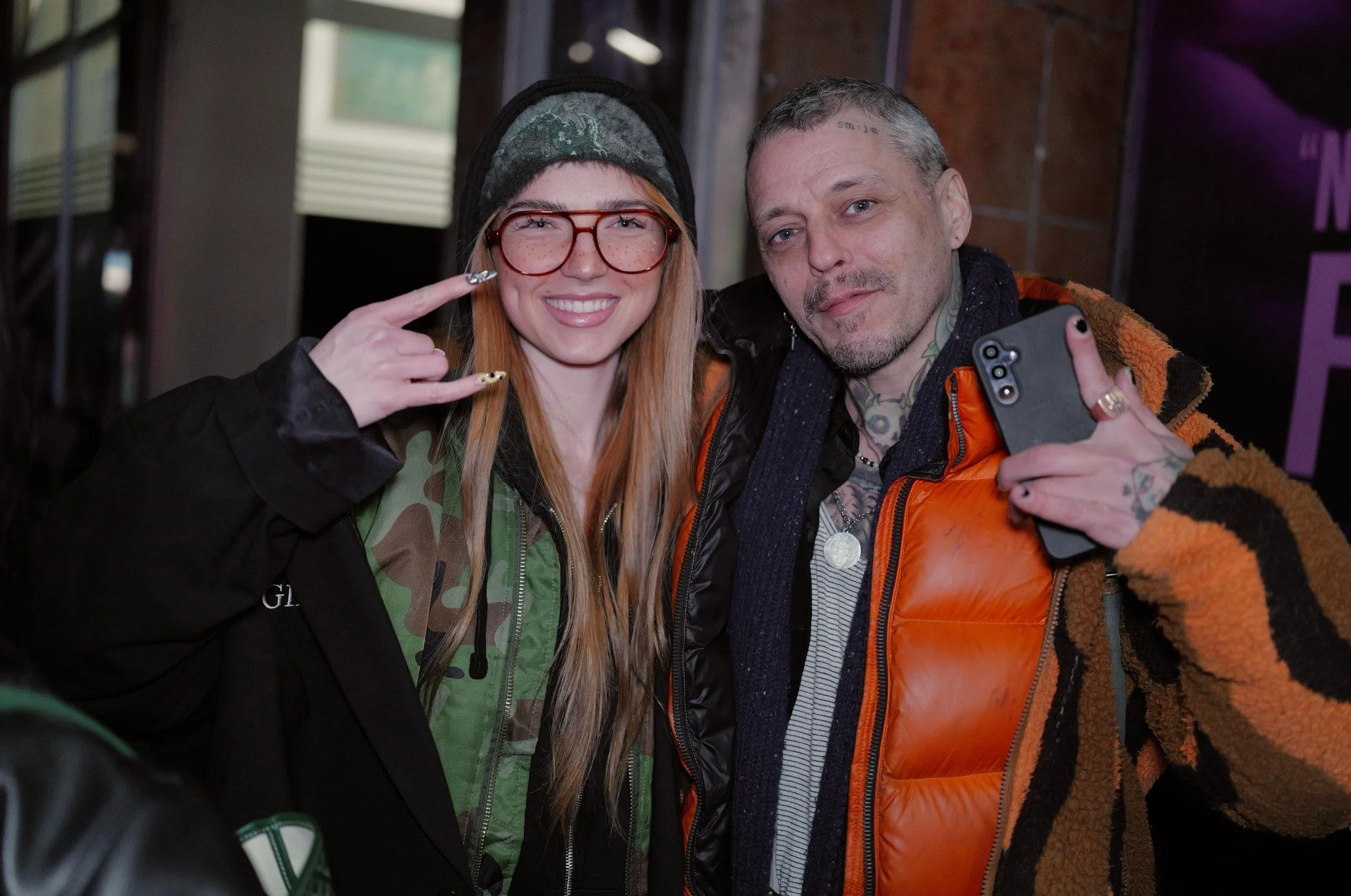 Two people posing, smiling; one wearing glasses and a camouflage jacket, the other in an orange jacket holding a phone.