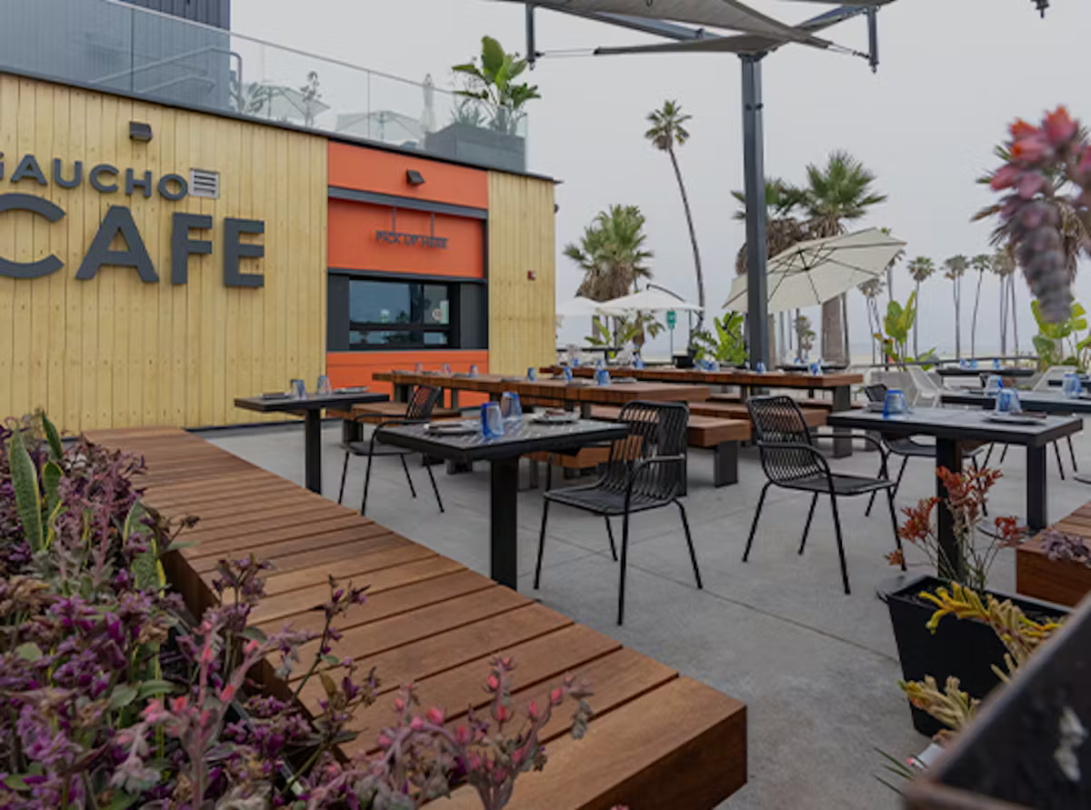 Outdoor cafe seating area with wooden tables and chairs, branded sign saying 'Gaucho Cafe,' potted plants, and umbrellas, with palm trees in the background.