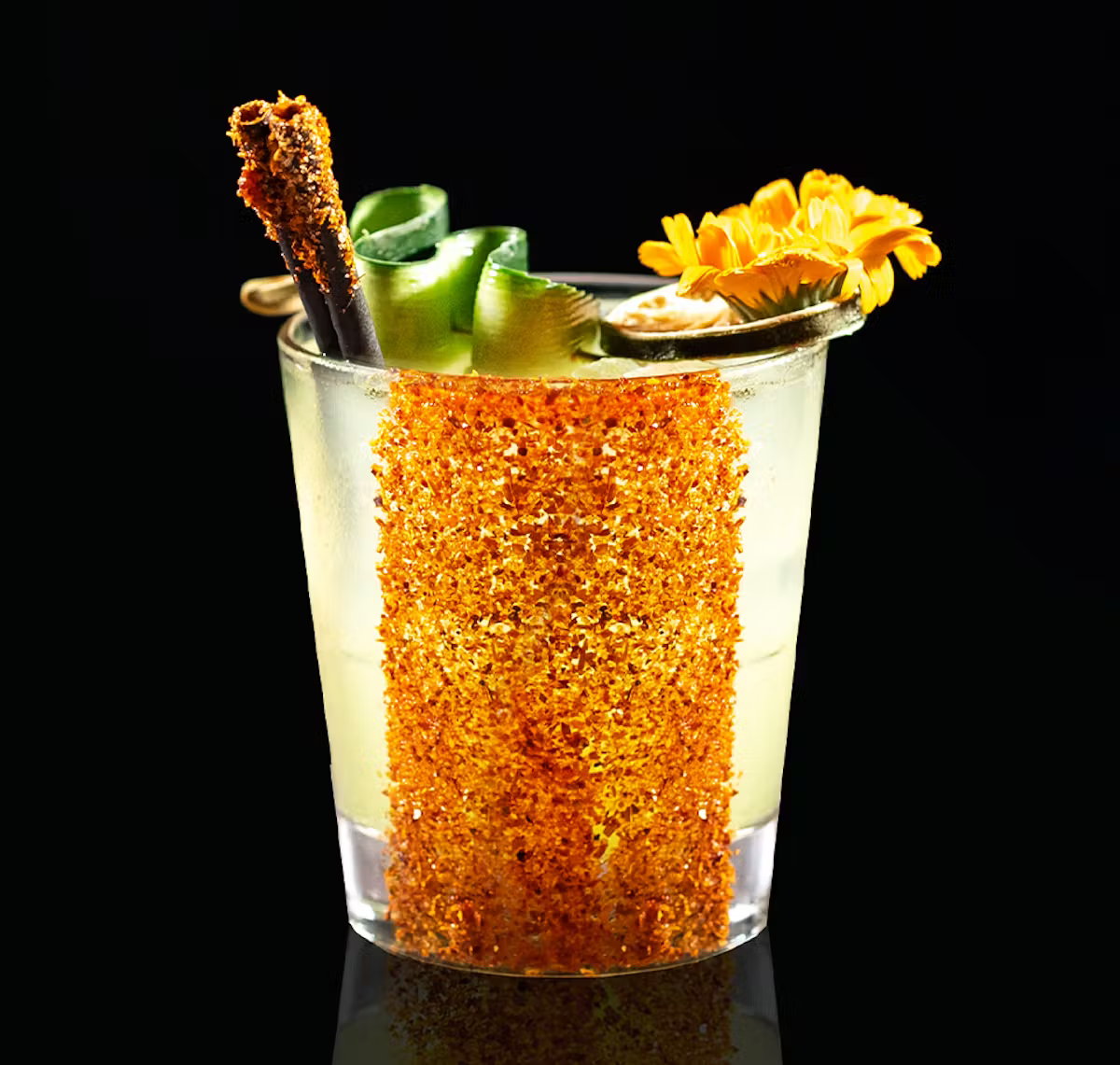 Cocktail in a glass with a spicy rim, garnished with cucumber spirals, a cinnamon stick, and an orange flower on a black background.