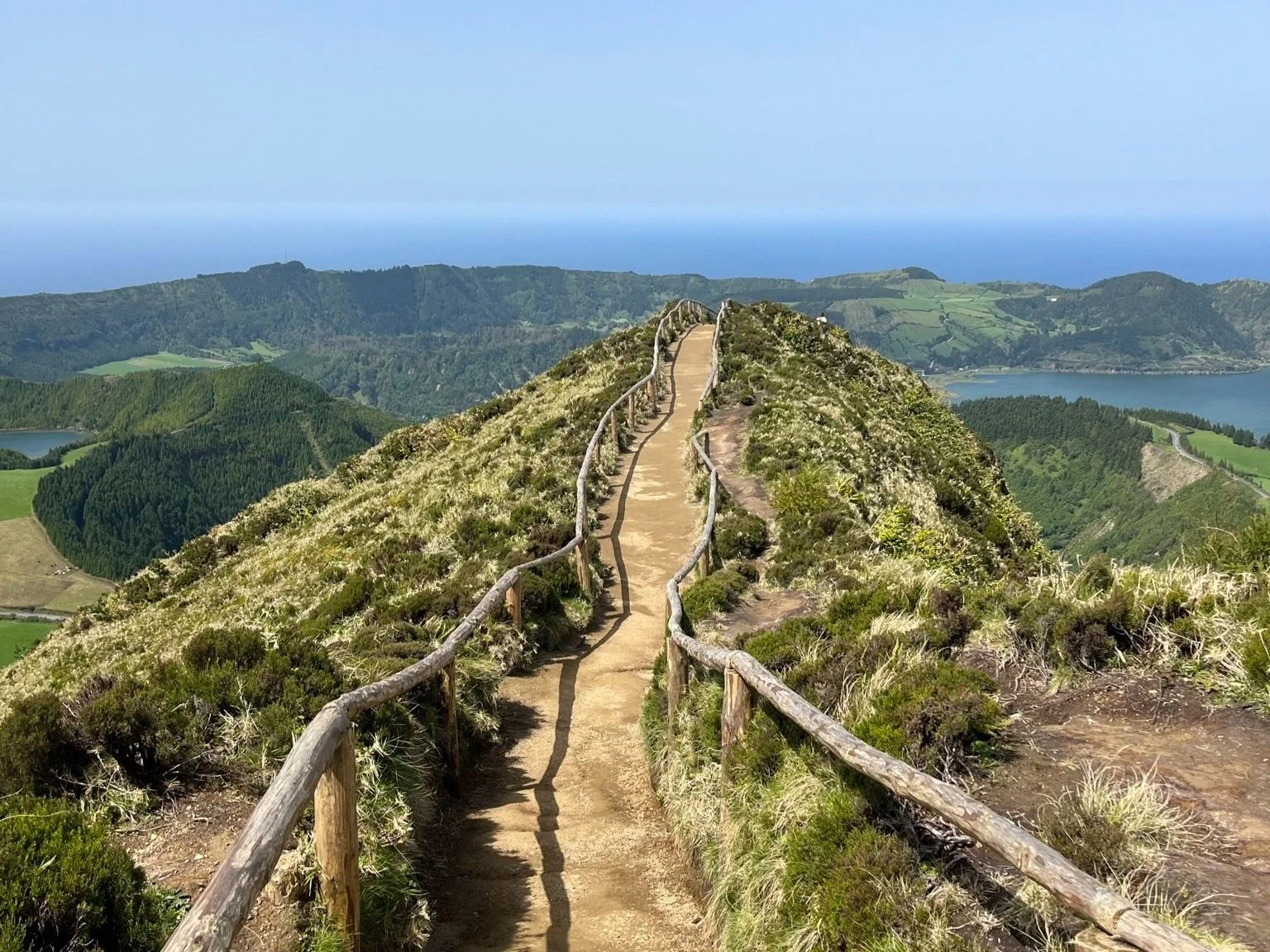 Scenic hiking trail with wooden railing in the Azores overlooking lush green landscape and blue ocean on a clear day.