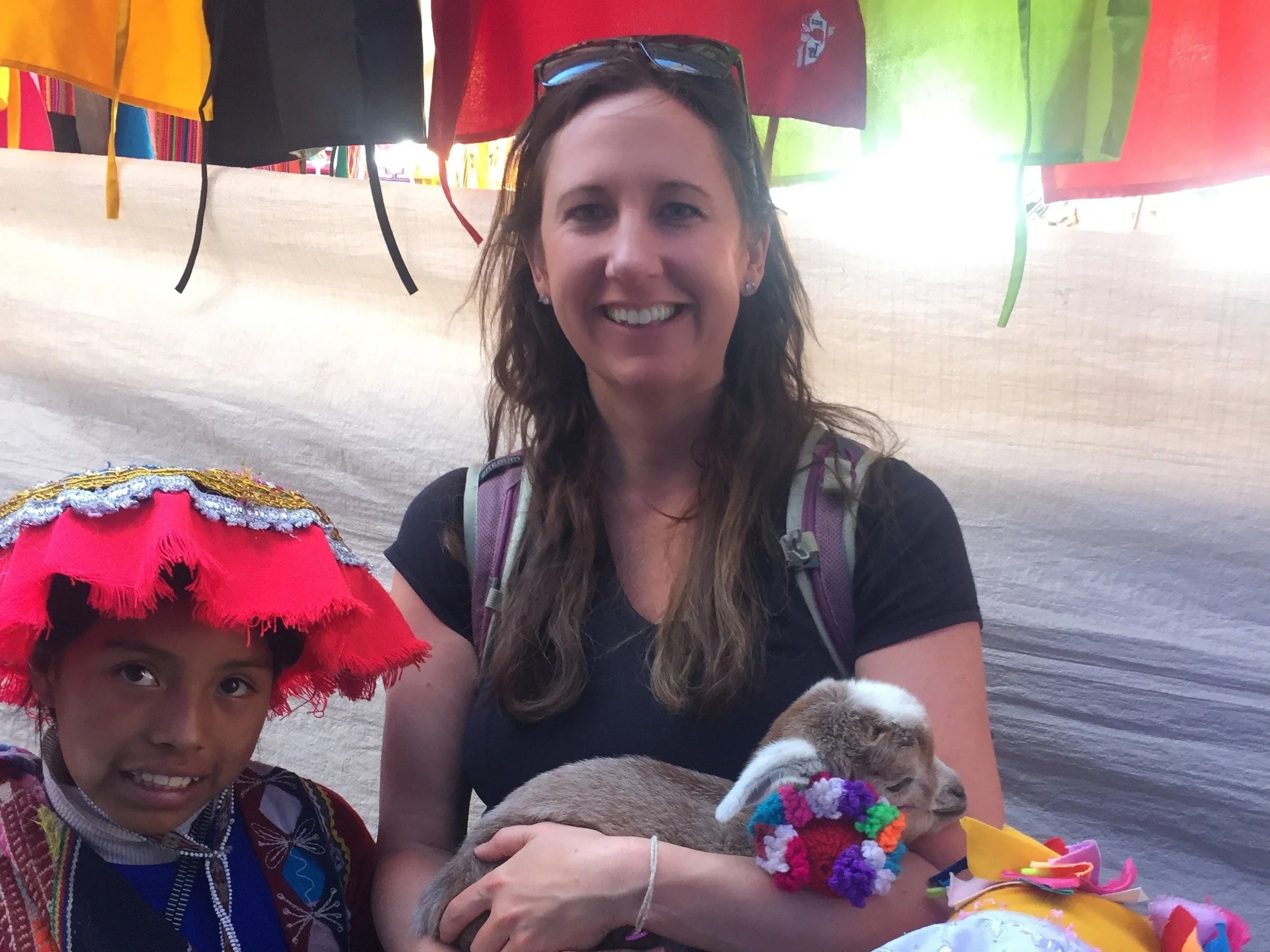 A woman standing next to a Peruvian child in colorful traditional attire, holding a small lamb also adorned with bright decorations. They are in front of a backdrop of various textile colors.
