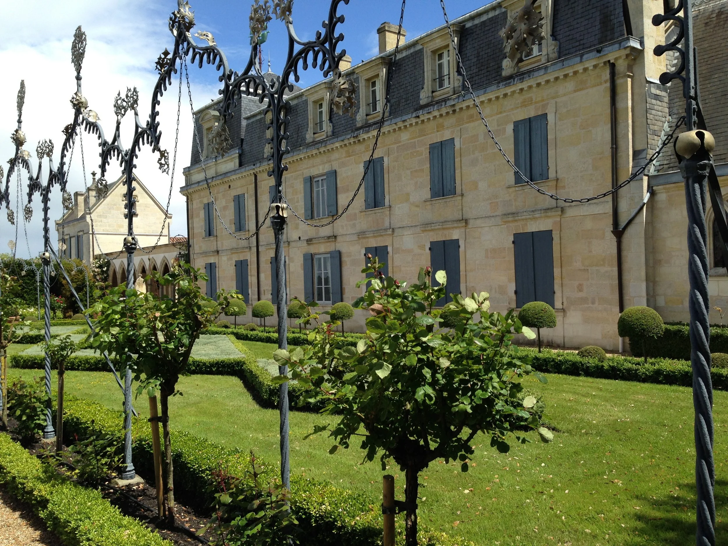 French historic stone building with blue shutters, ornate iron fencing, and manicured garden with topiary shrubs on a sunny day.