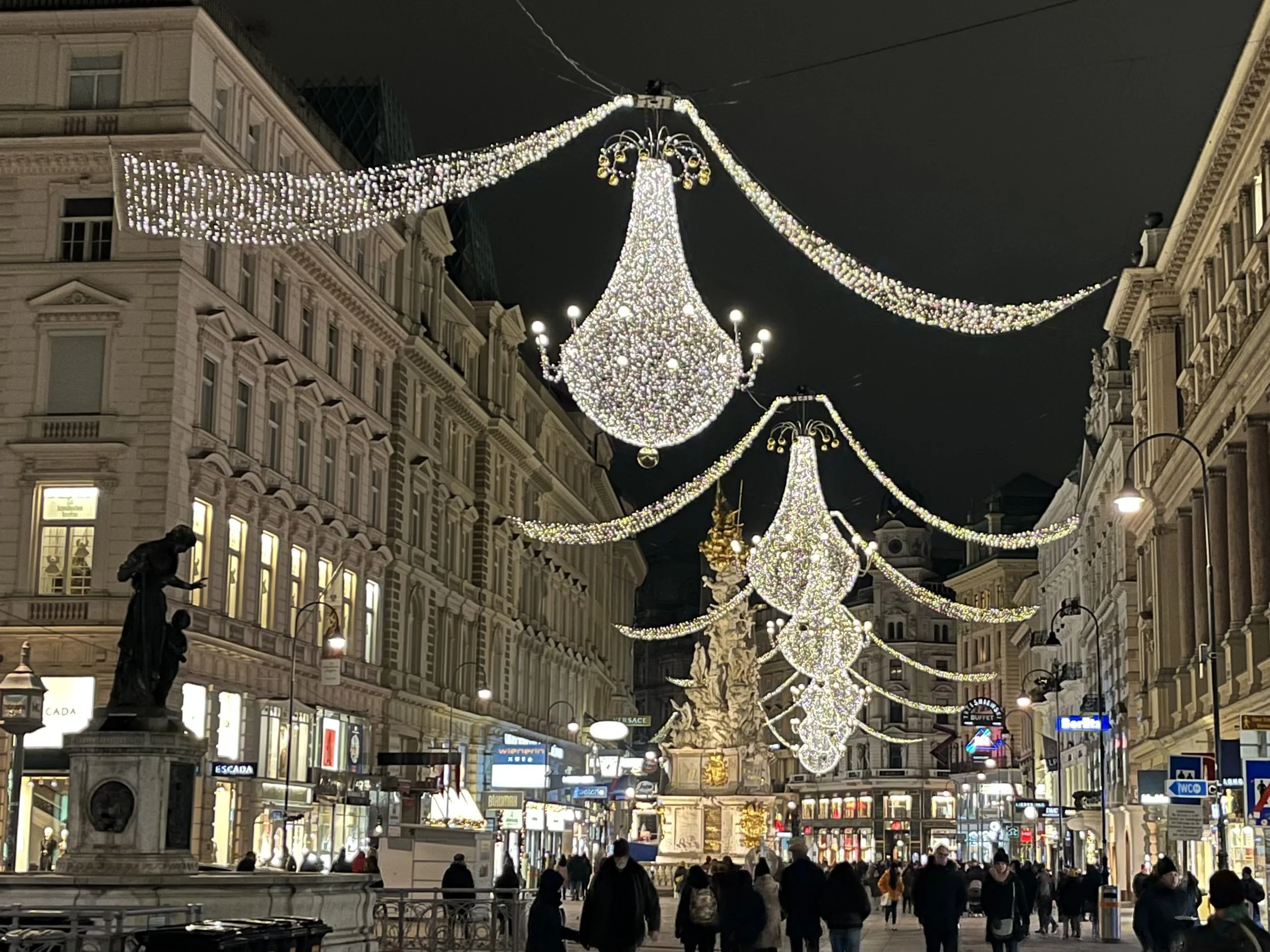 A city street at night, decorated with large, elegant chandelier-like lights hanging overhead. The street is lined with historic buildings and shops, with people walking along the sidewalk. The area is brightly lit, creating a festive and welcoming atmosphere.
