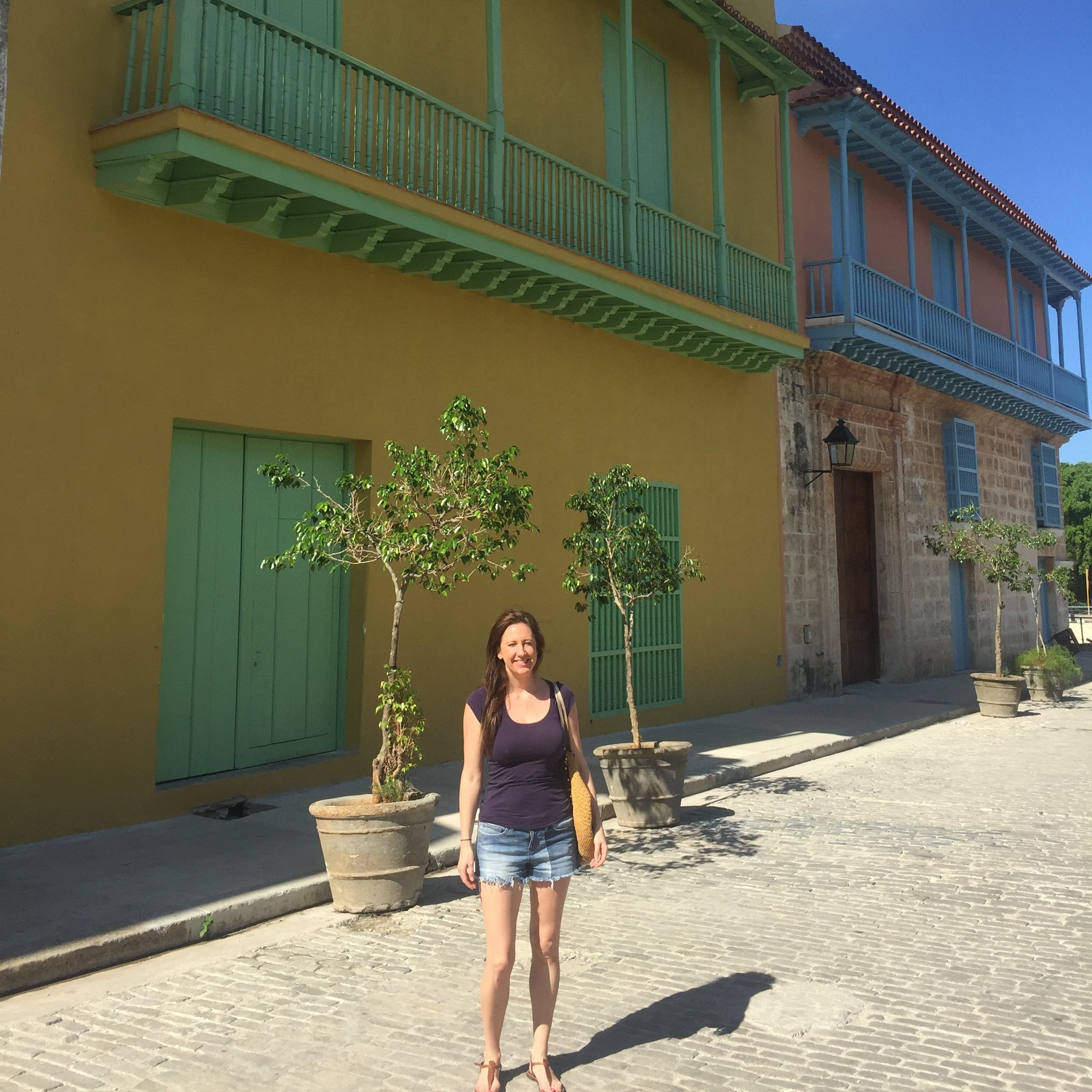 Woman in casual outfit standing on cobblestone street of Cuba in front of colorful colonial-style buildings with potted plants and bright blue sky.
