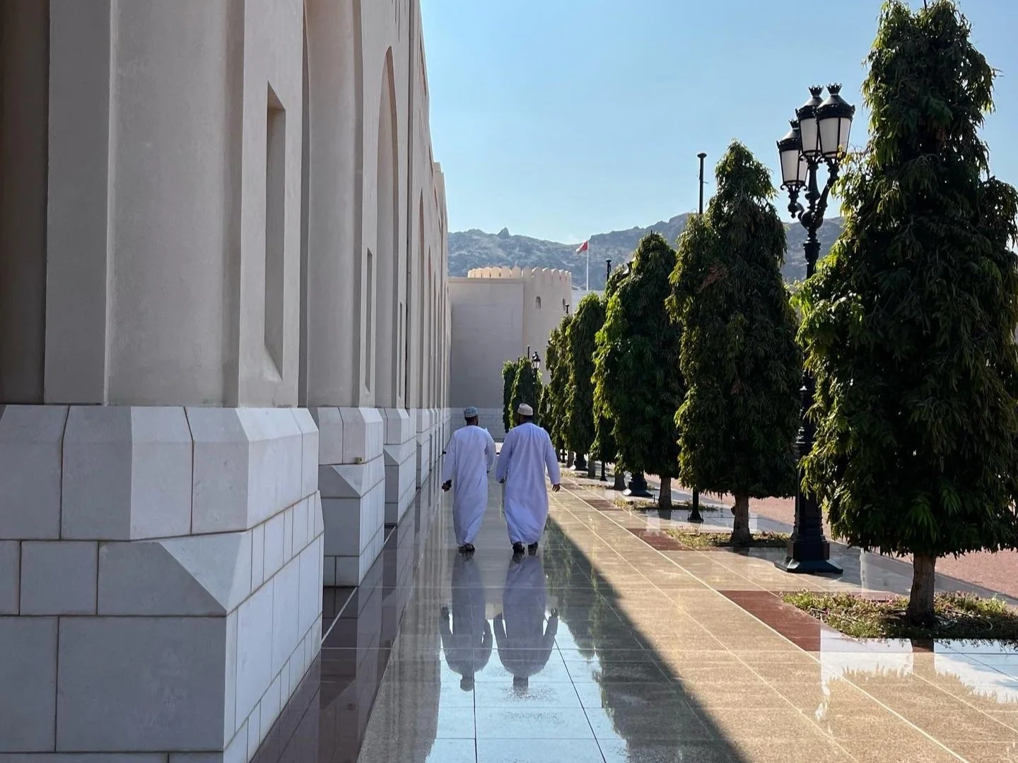 Two people wearing traditional white clothing in Oman walking along a street lined with neatly trimmed trees and ornate lamp posts, adjacent to a large cream-colored building.
