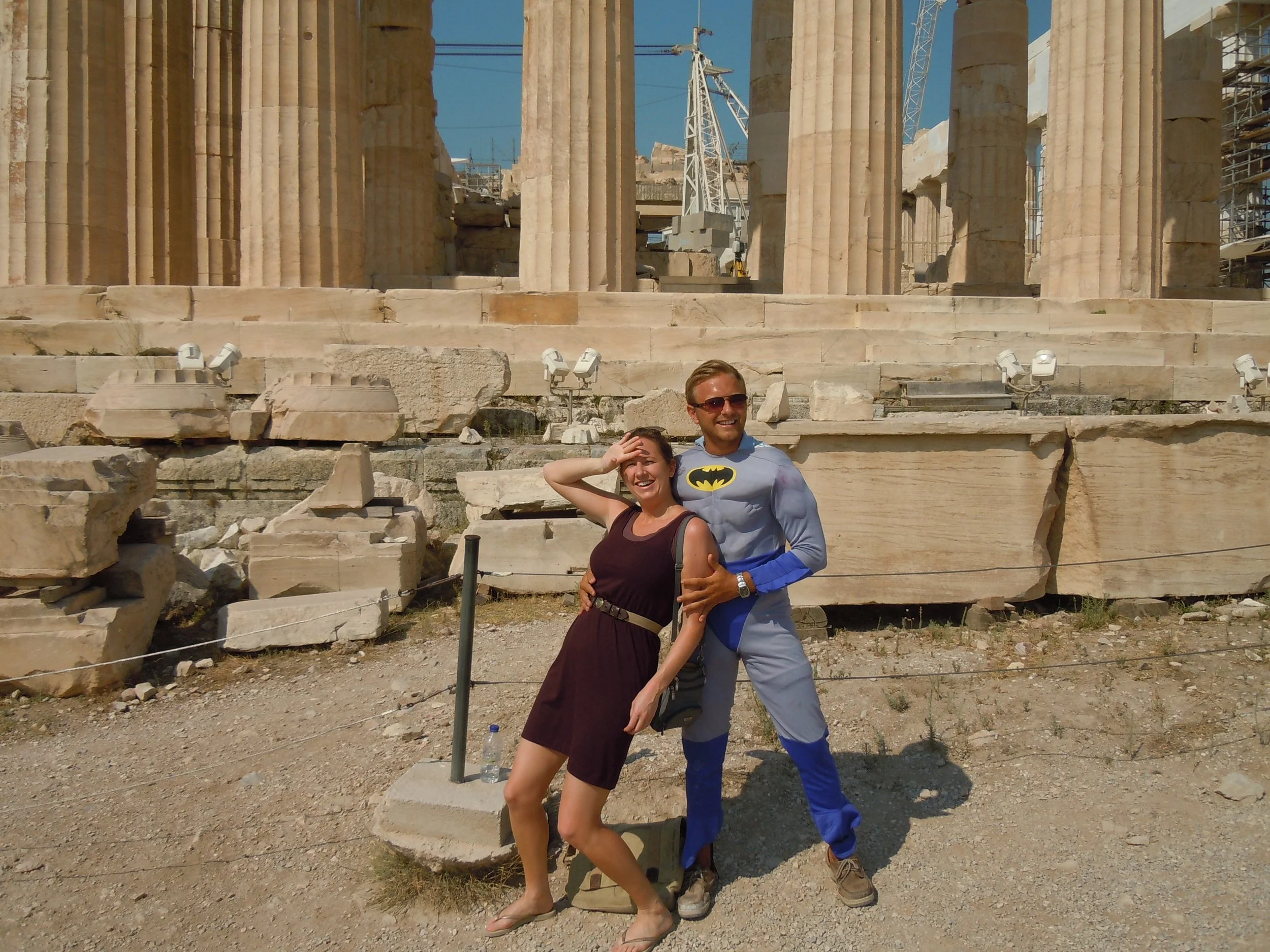 Two people posing in front of ancient Greek columns; one wearing a superhero costume, the other in a dress.