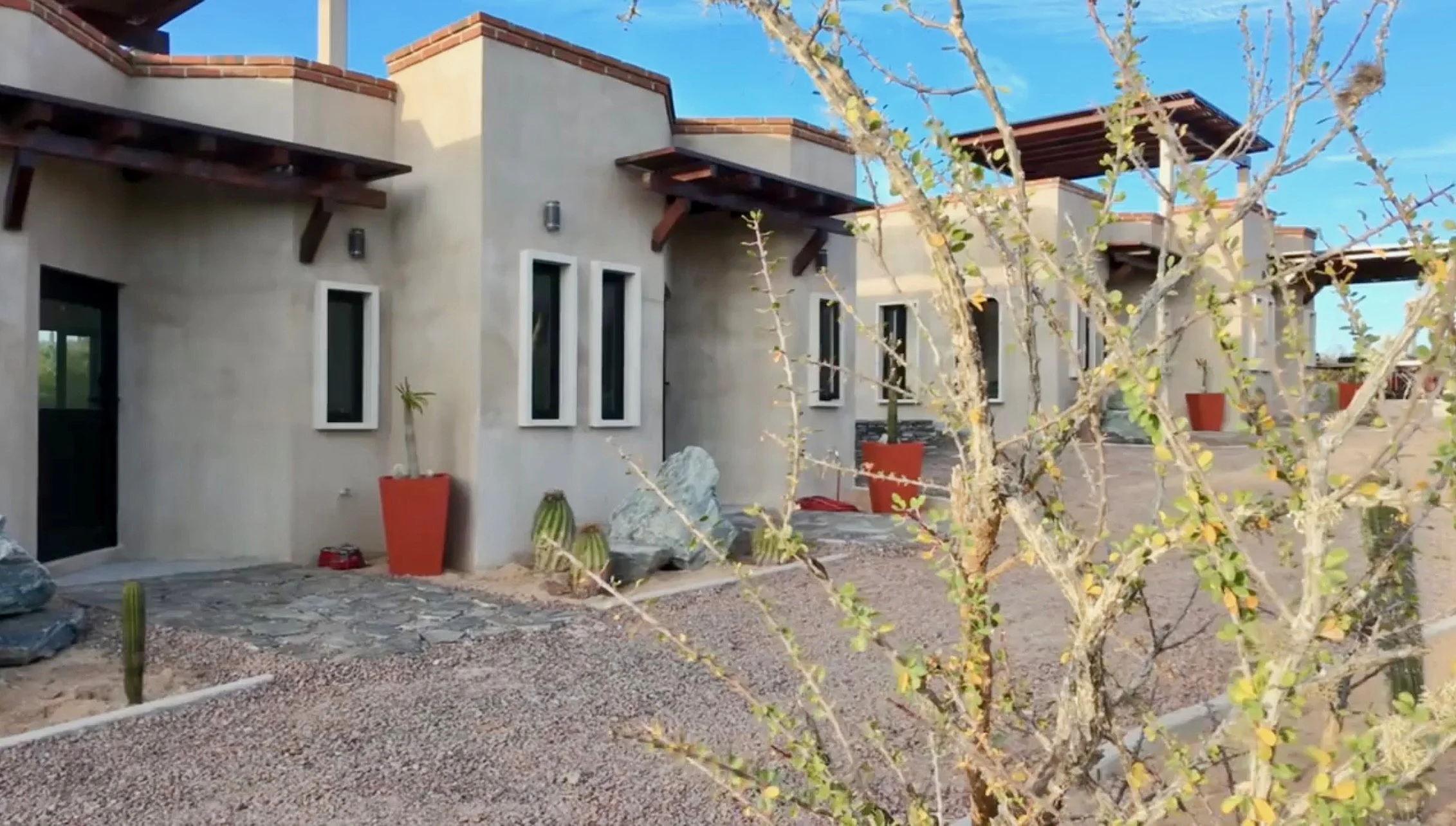 Exterior of a modern artist studio with beige stucco walls, multiple narrow windows, and wooden awnings. The yard features desert landscaping with gravel, cacti, and a leafless bush in the foreground under a blue sky.