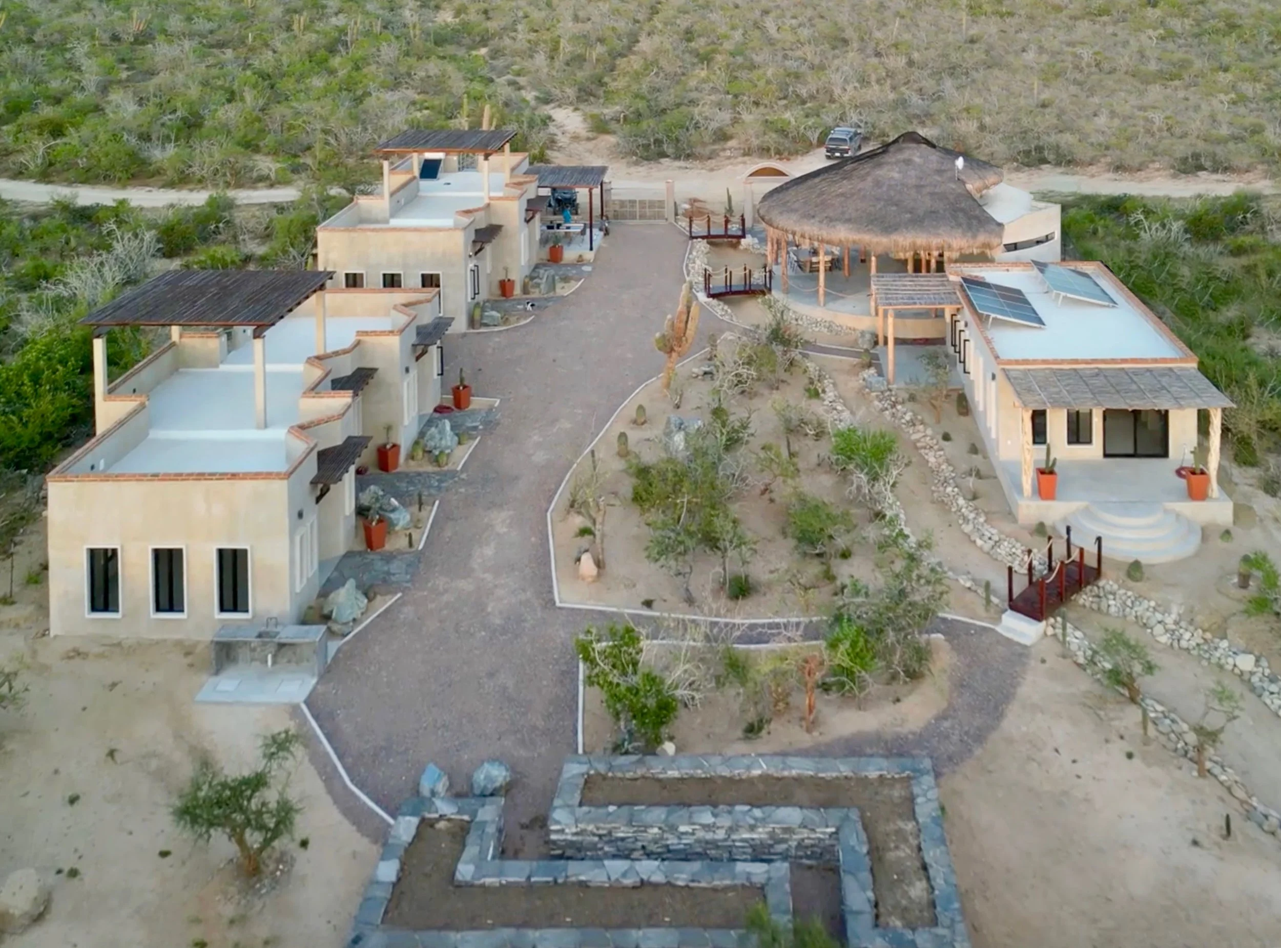 Aerial view of a commercial property with multiple modern buildings, a thatched-roof structure, a landscaped garden with trees, and solar panels on some roofs, set in a desert environment.