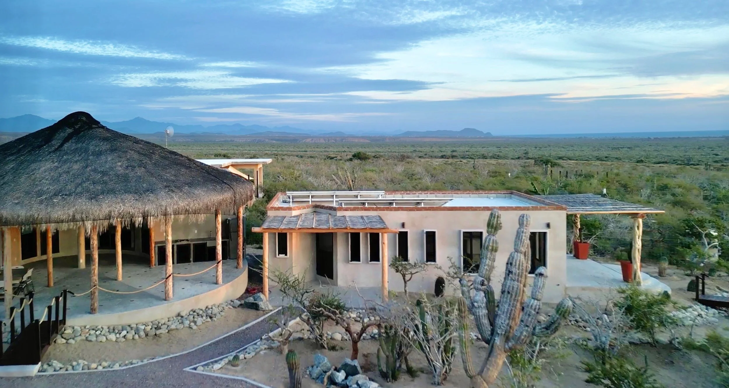 Scenic view of a desert landscape with two modern buildings, one with a thatched roof, surrounded by cactus plants and dry vegetation under a cloudy sky.