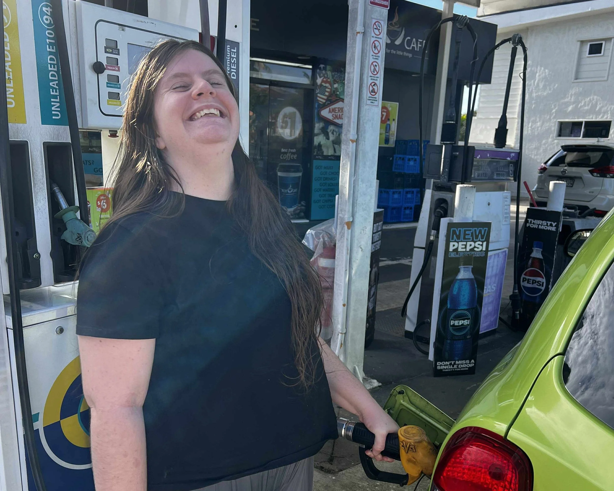A woman with long brown hair and a black shirt is smiling while filling up her yellow-green car with gas at a gas station, with gas pumps and signage visible in the background.