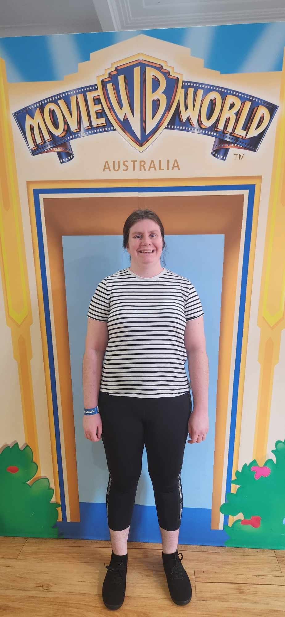 A woman standing in front of a colorful sign that says 'Warner Bros Movie World Australia' with a cartoon-style frame around her.