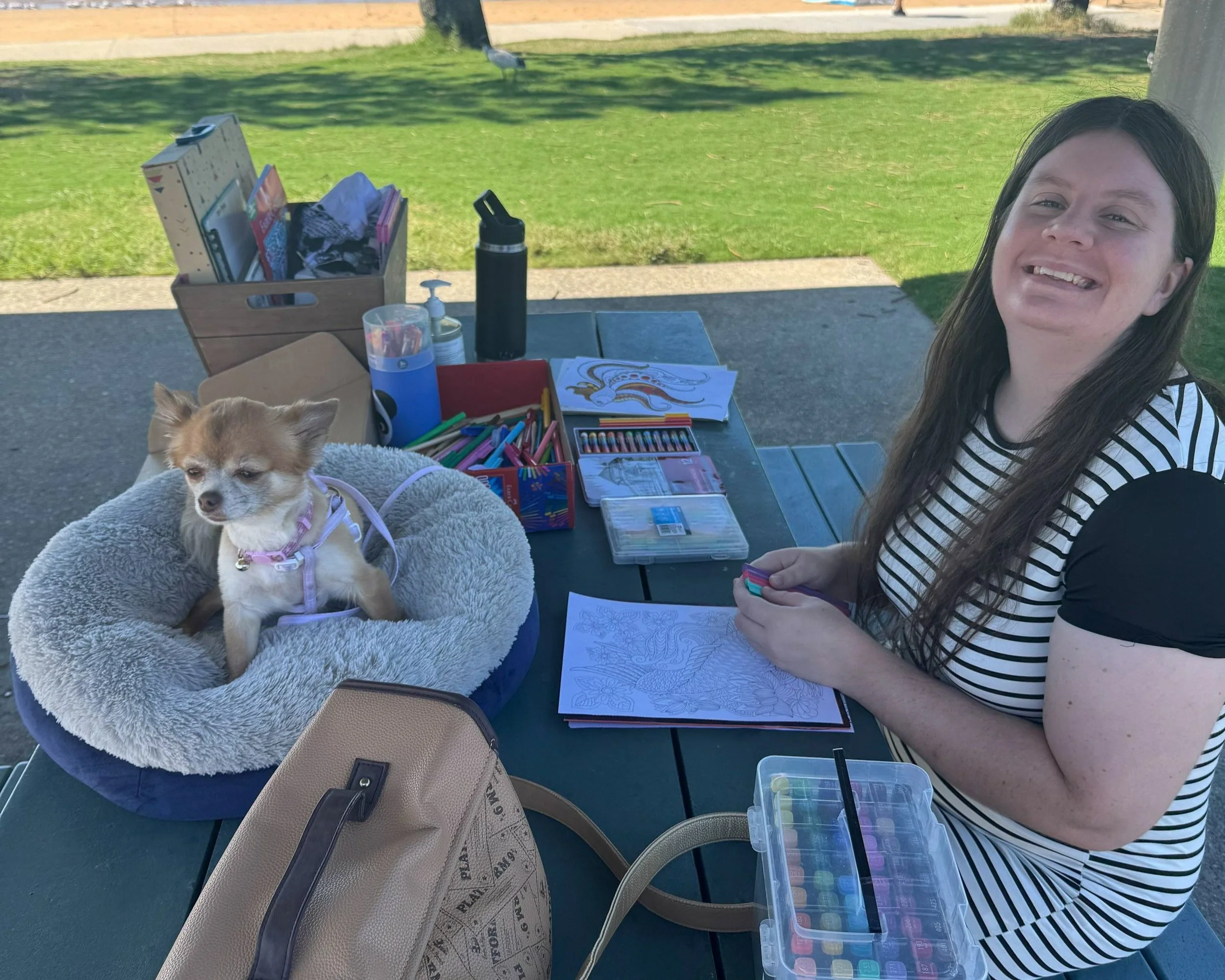 A woman with long brown hair smiling at a picnic table outdoors, with a small dog in a cozy bed next to her. The table has coloring supplies, markers, and coloring pages. Grass and trees are in the background.