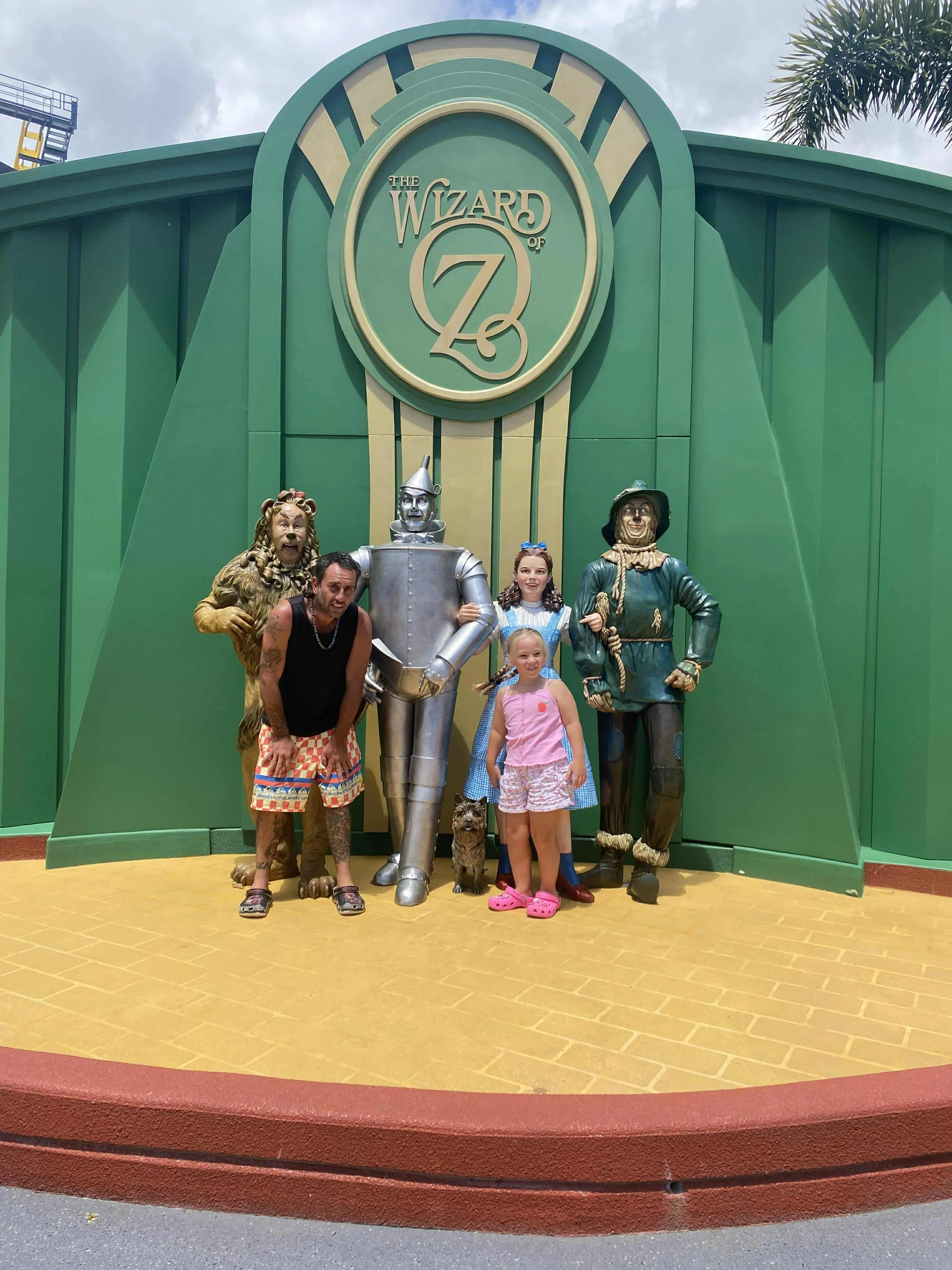 Family posing in front of a green sign with statues of characters from The Wizard of Oz, including the Cowardly Lion, Tin Man, Dorothy, and Scarecrow.