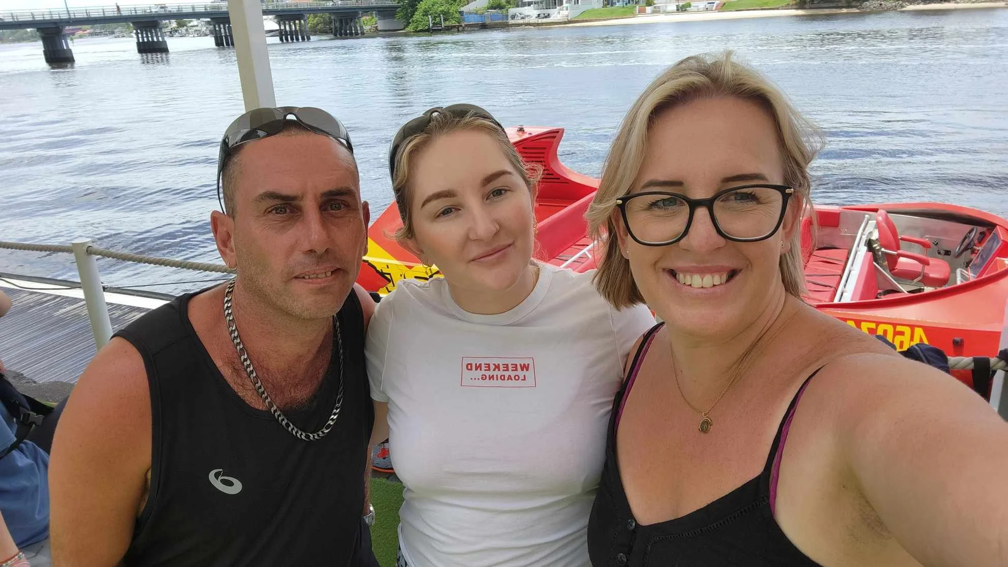 Three people taking a selfie near a body of water with two red boats in the background.