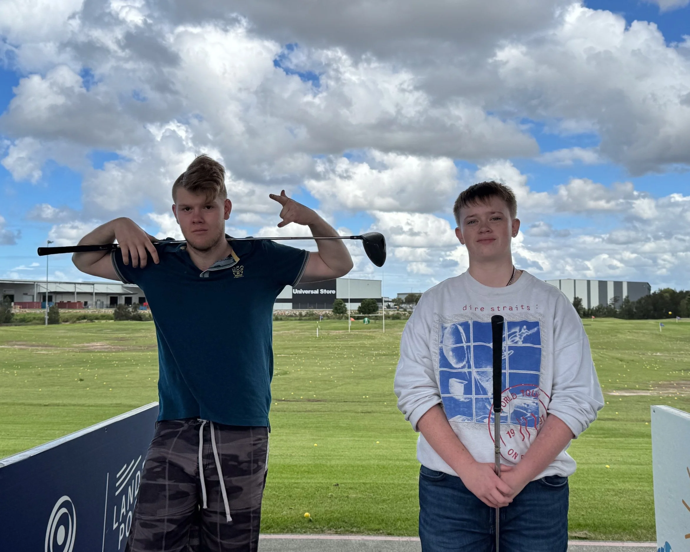 Two young boys stand on a golf driving range with a grassy field and a large building in the background. One boy, on the left, is holding a golf club across his shoulders and making a peace sign with his right hand. The other boy, on the right, is holding a golf club vertically and smiling. The sky is partly cloudy.