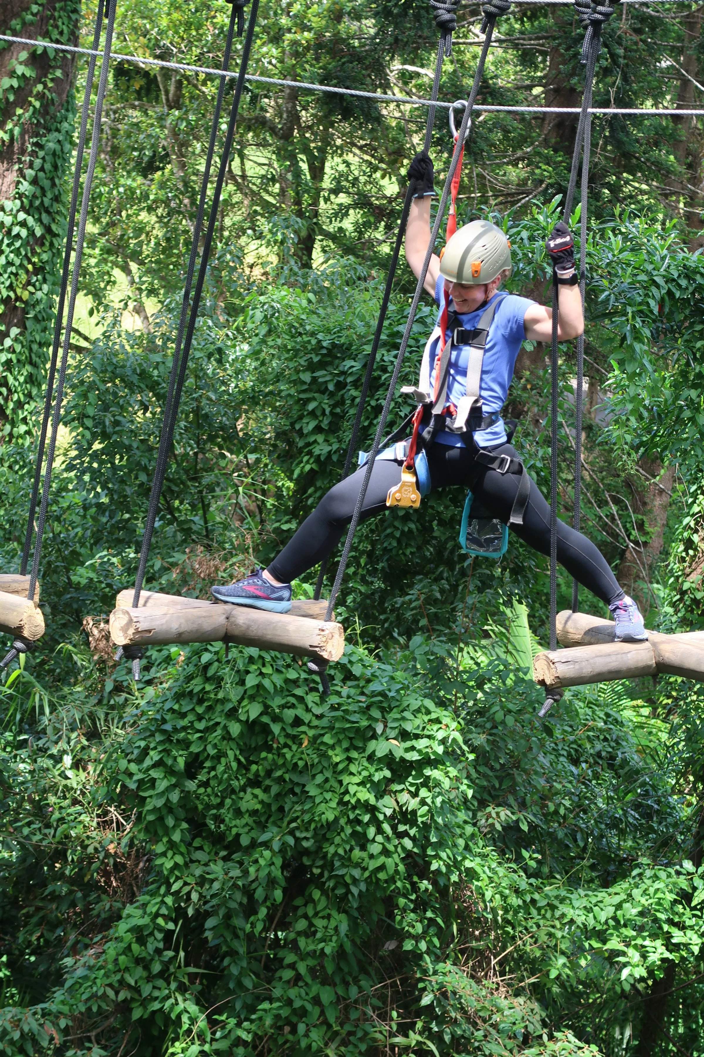 A young girl in a helmet and harness crossing a ropes course on a wooden bridge in a dense green forest.