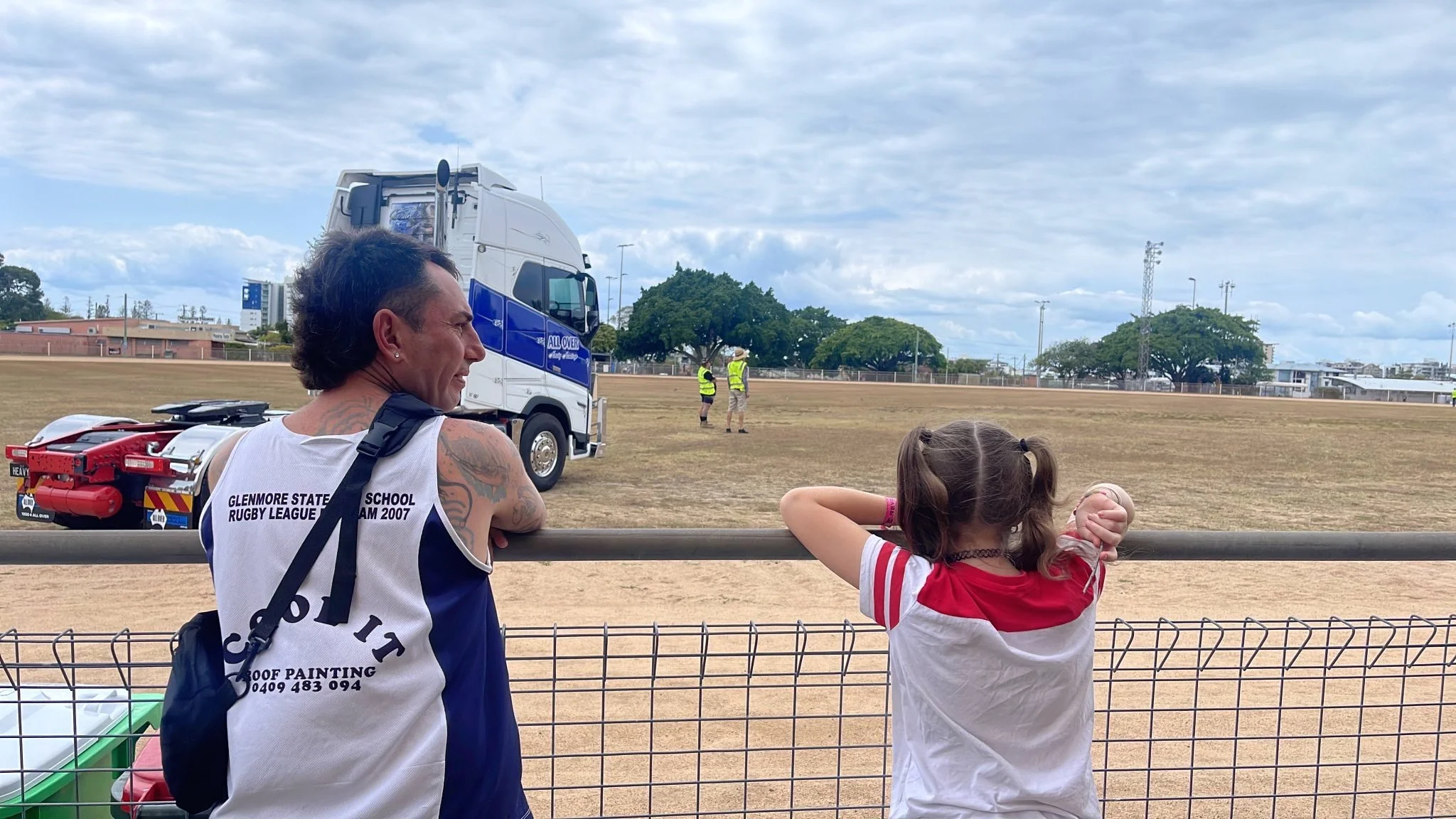 A man and a young girl are watching a rugby practice or game on a field behind a metal fence. The man is wearing a sleeveless sports jersey and has tattoos, while the girl has pigtails and is wearing a red and white shirt. Two people in high-visibility vests are in the background on the field, and a large truck is parked nearby.